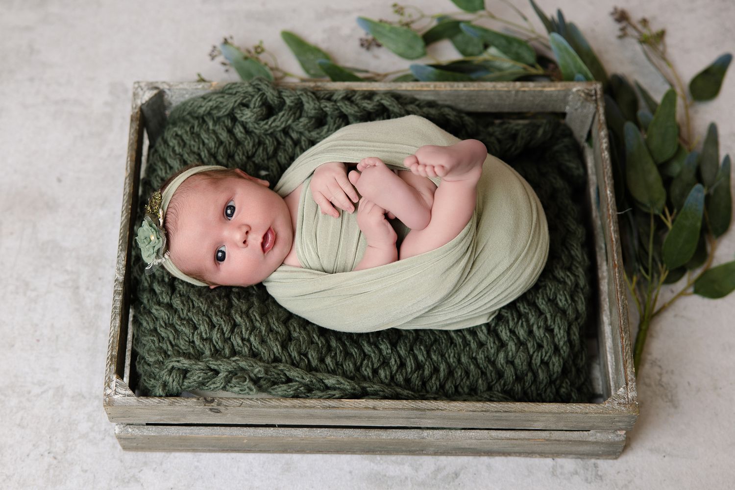 Newborn baby swaddled in sage green wrap lying in rustic wooden crate with knit blanket and eucalyptus.