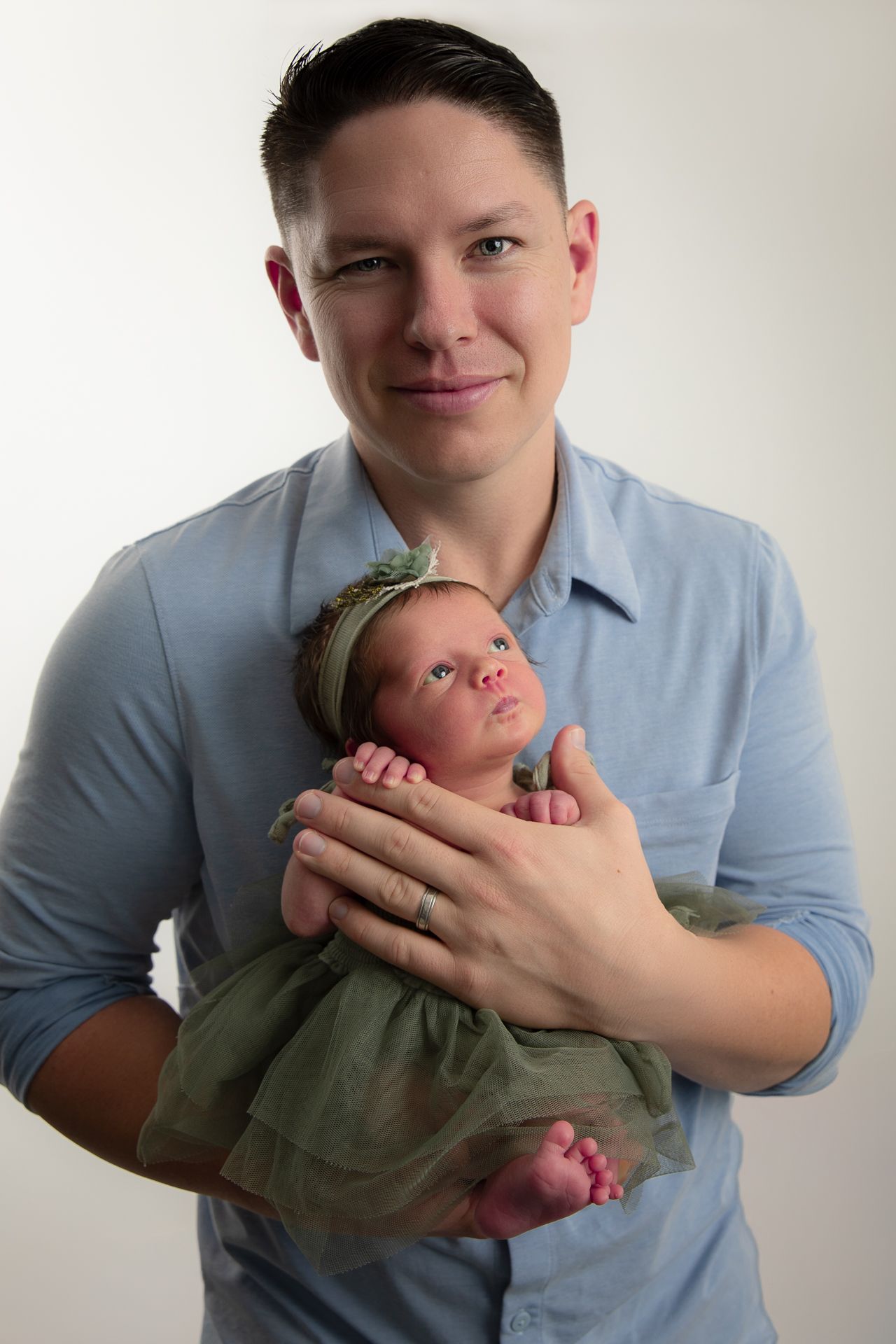 Parent in blue shirt holding a newborn baby dressed in green with a floral headband against white background.