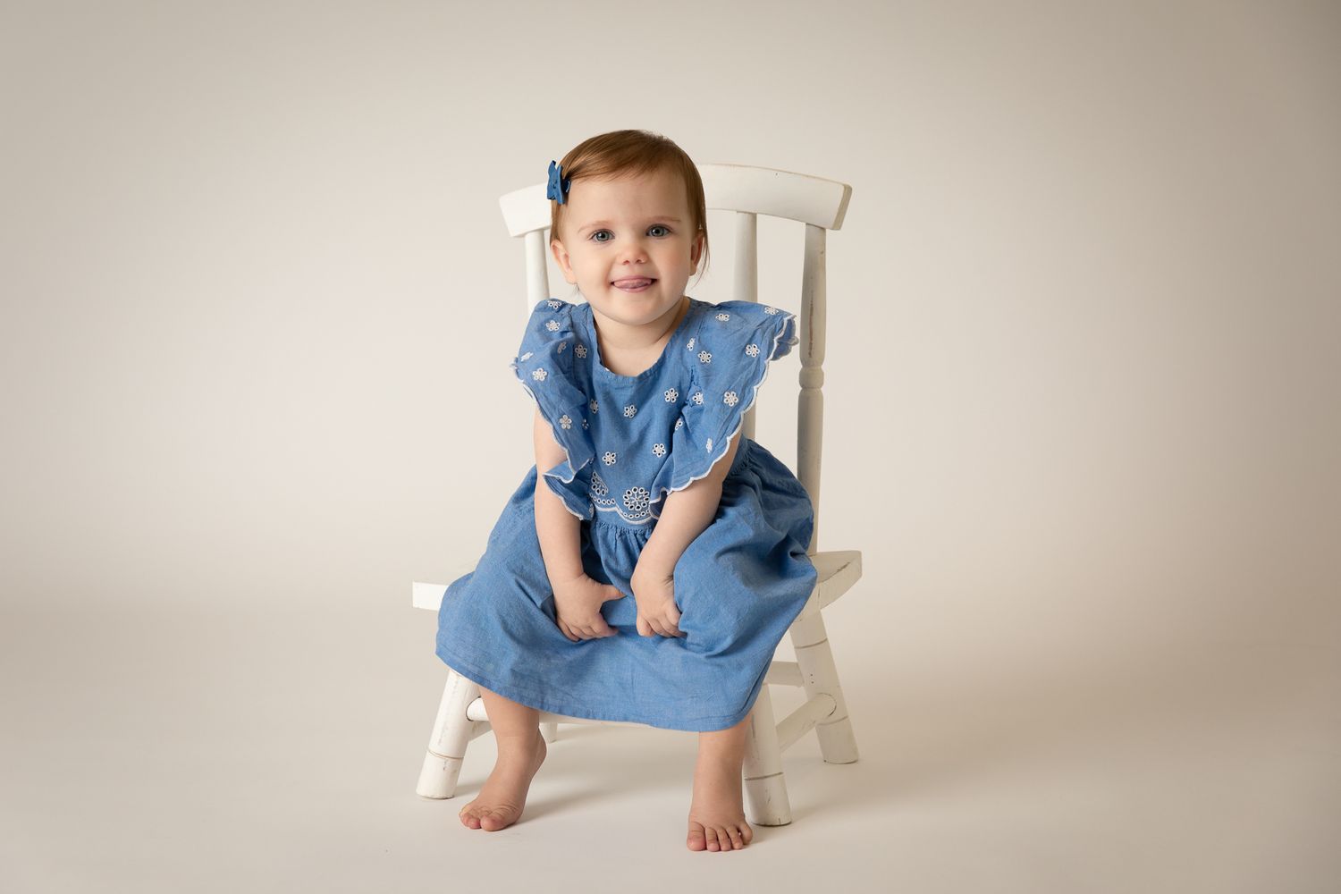 Smiling baby girl in blue polka dot dress sitting on white chair.