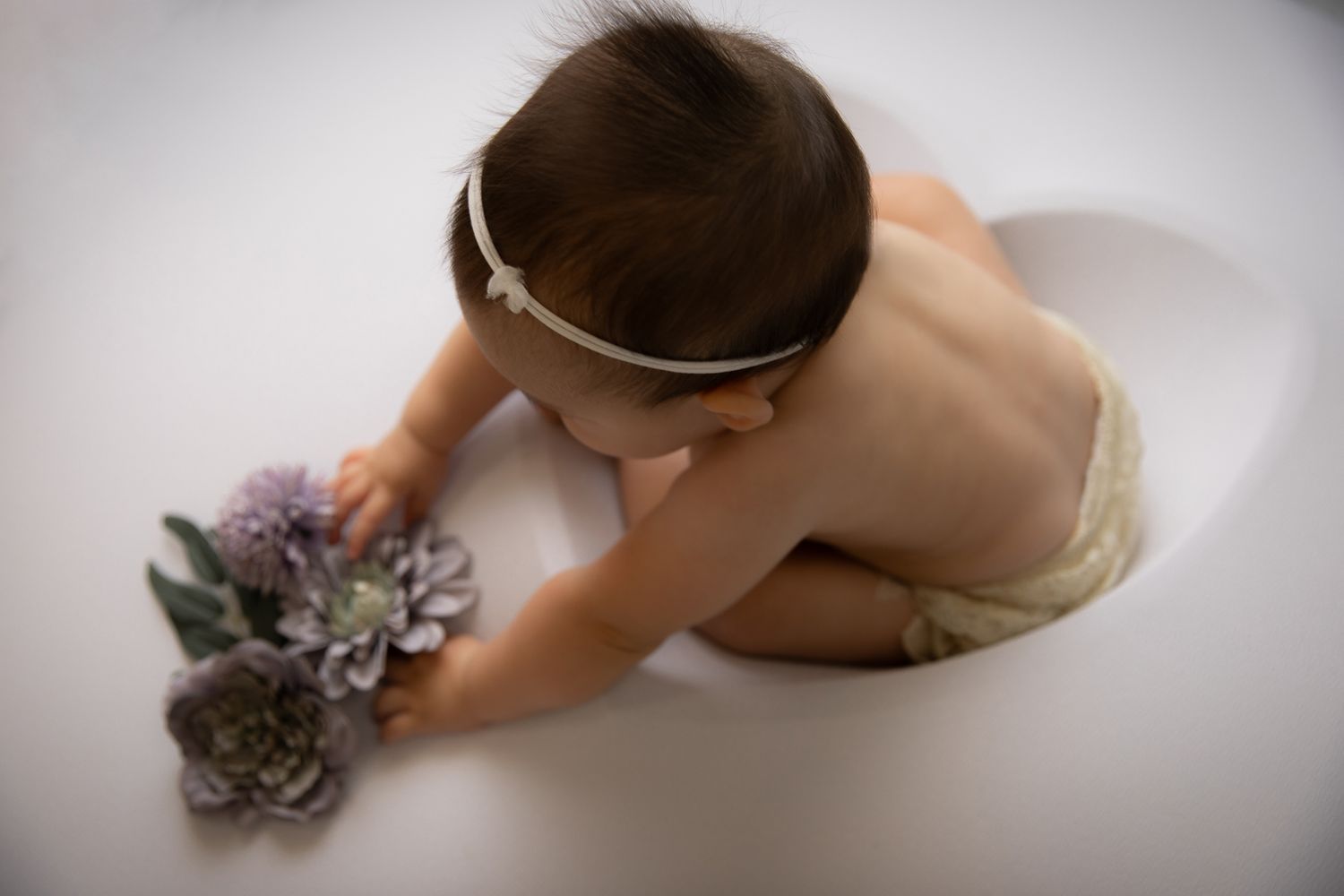 Baby girl in white headband reaching for purple flowers on white studio floor.