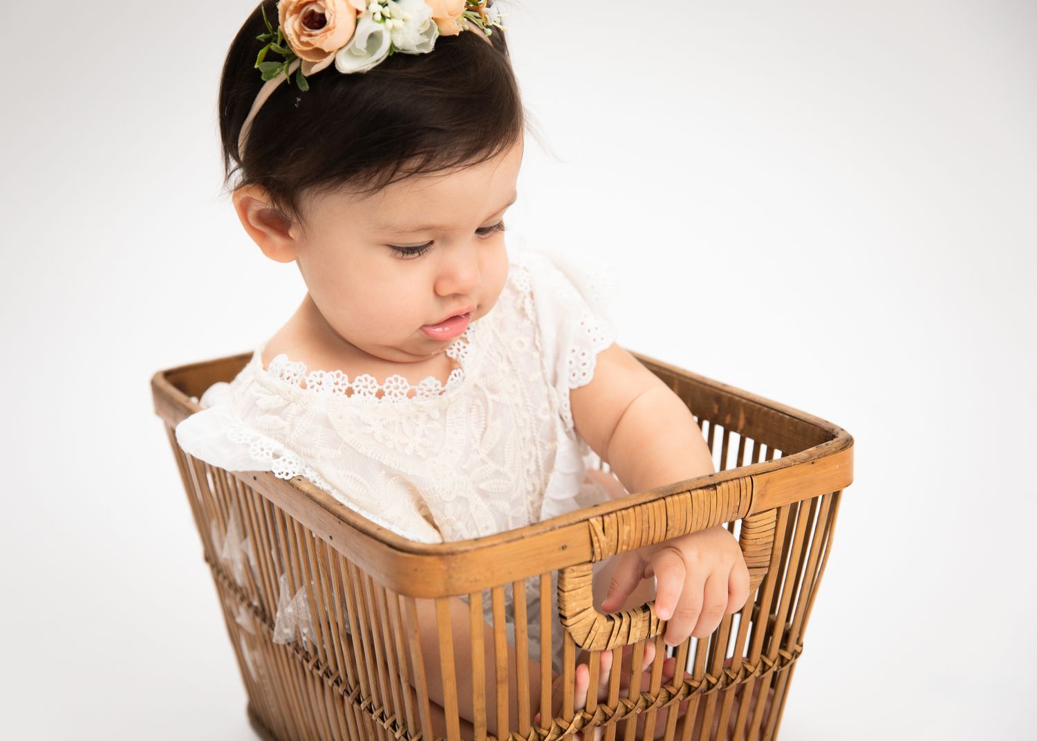 Adorable baby girl with floral headband looking down while sitting in a wooden basket.