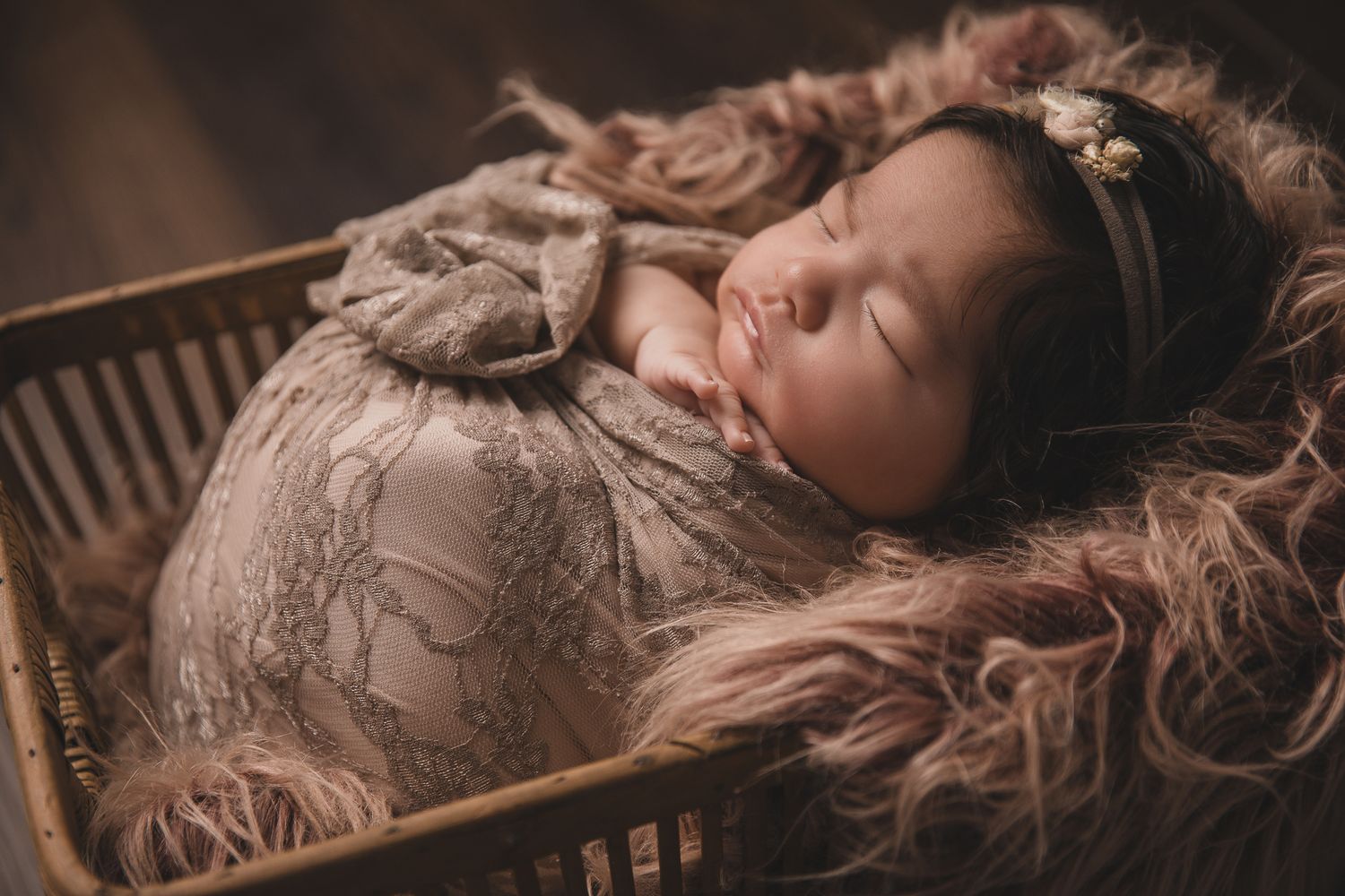 Sleeping newborn baby wrapped in lace swaddle with floral headband in wooden basket with fluffy fur.
