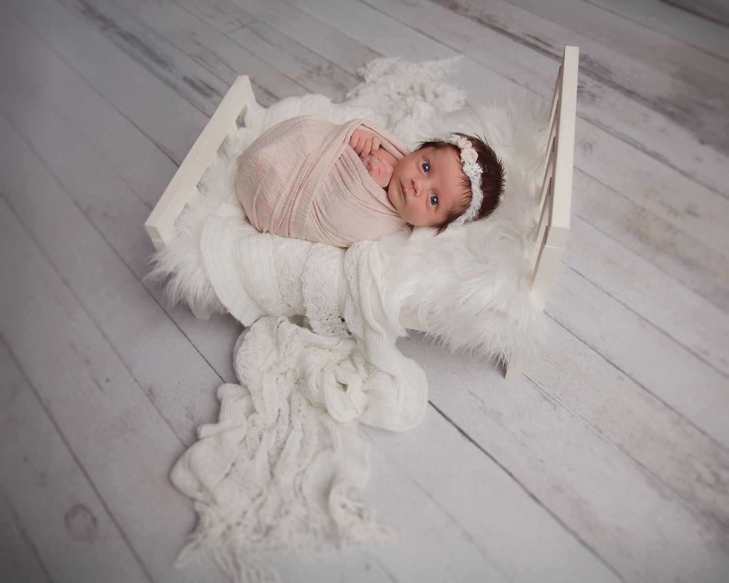 Awake newborn baby girl in pink wrap with crystal headband lying in white mini bed prop. Vista photographer