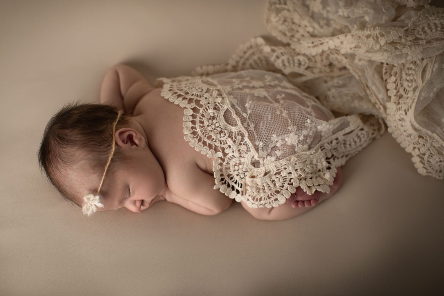 Sleeping newborn baby wearing lace romper and floral headband on neutral background.