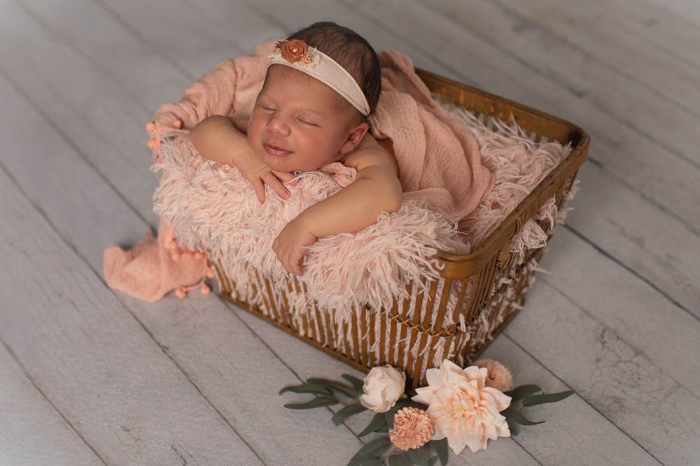 A sleeping newborn in pink clothing rests peacefully in a rustic wooden basket with floral decorations on a gray floor.
