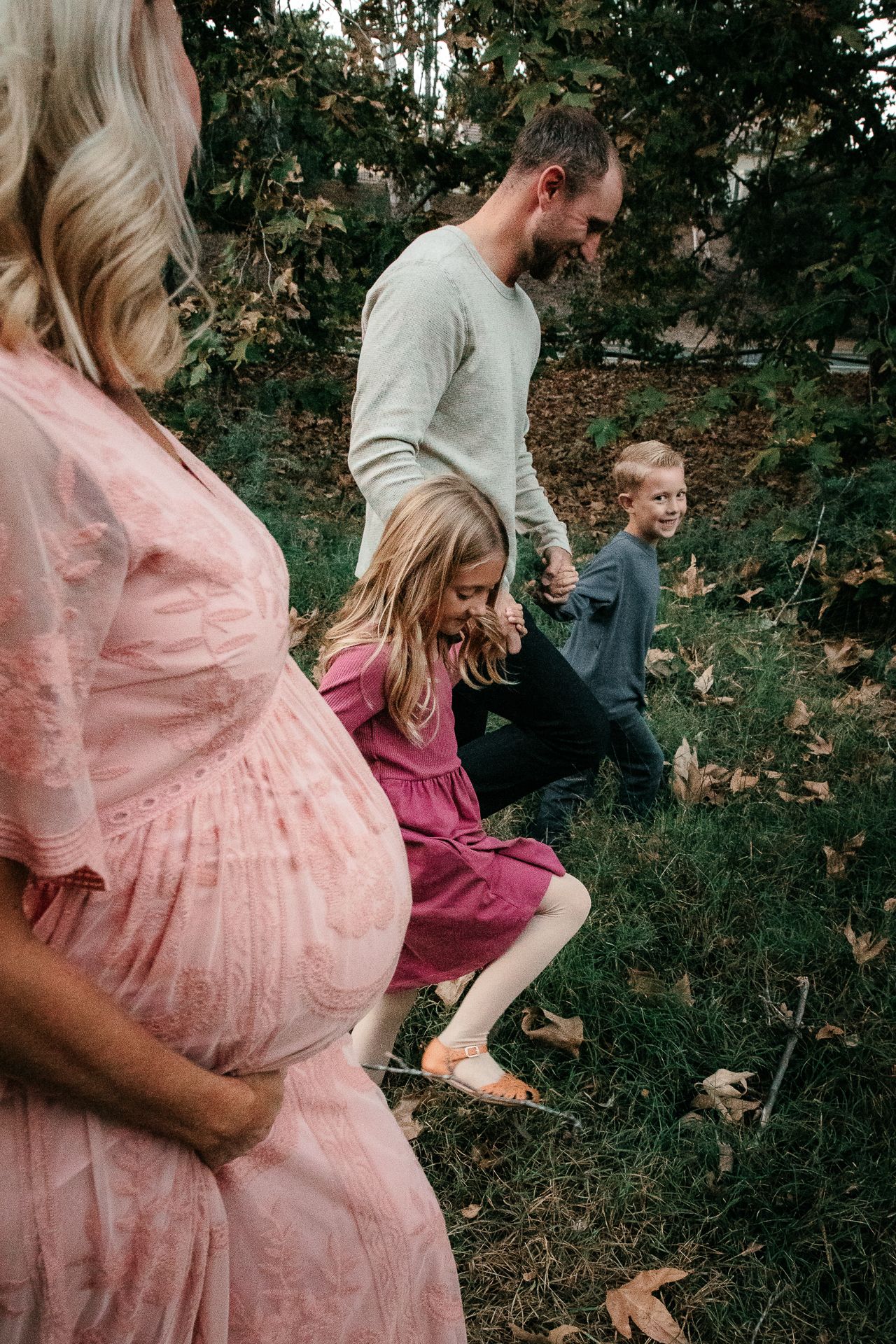A pregnant woman in pink dress with family walking outdoors among autumn leaves and green trees at a Fallbrook Preserve