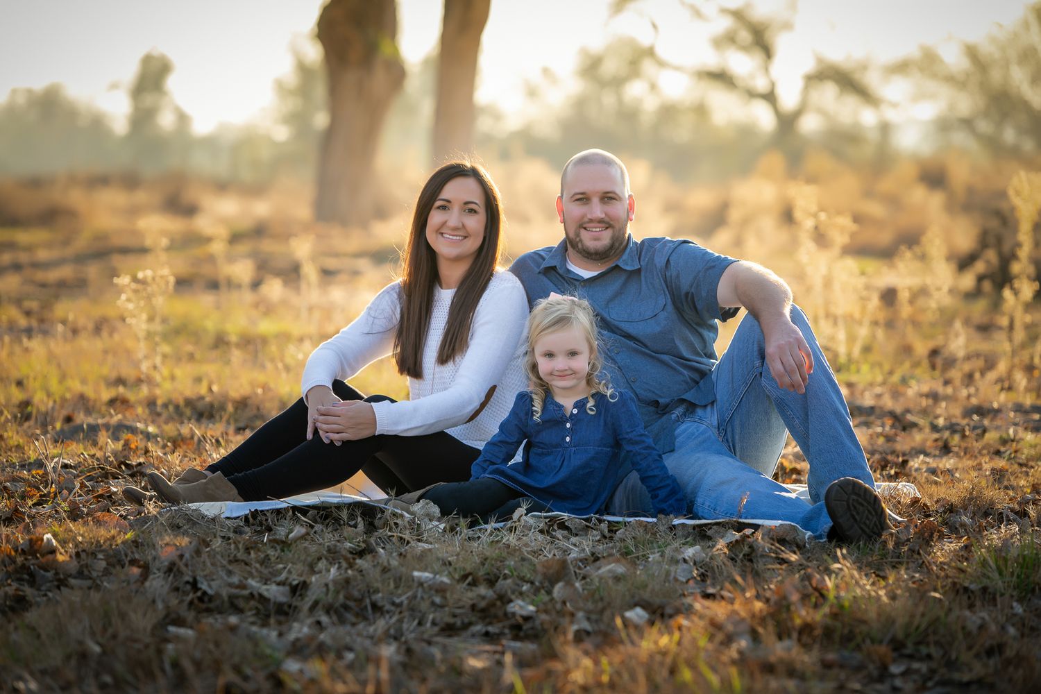 A family of three sits relaxed on a blanket in a sun-drenched autumn outdoor setting.