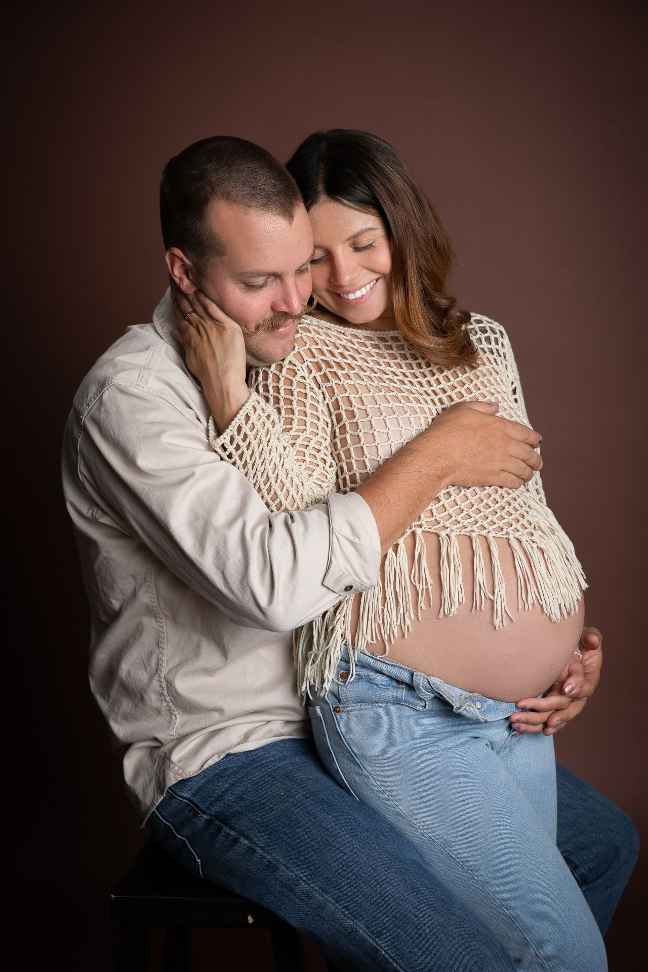 Smiling couple embracing in studio, pregnant woman in crochet poncho and jeans with partner holding her bump.
