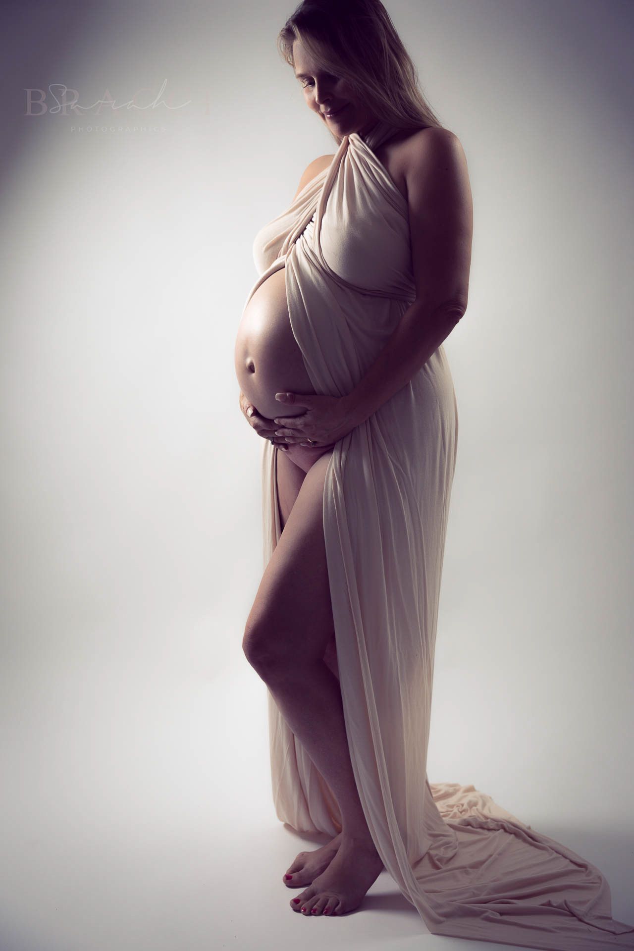 Pregnant woman draped in sheer fabric elegantly cradling her baby bump in soft light.