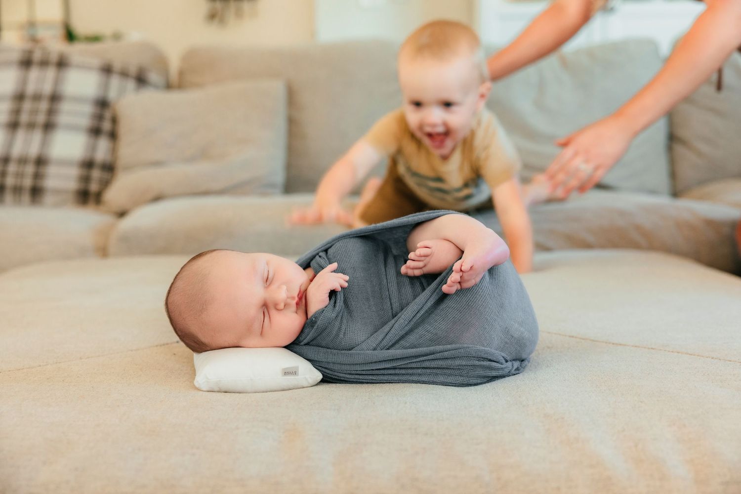 Newborn baby swaddled in gray wrap sleeping on couch while excited toddler sibling plays in background.