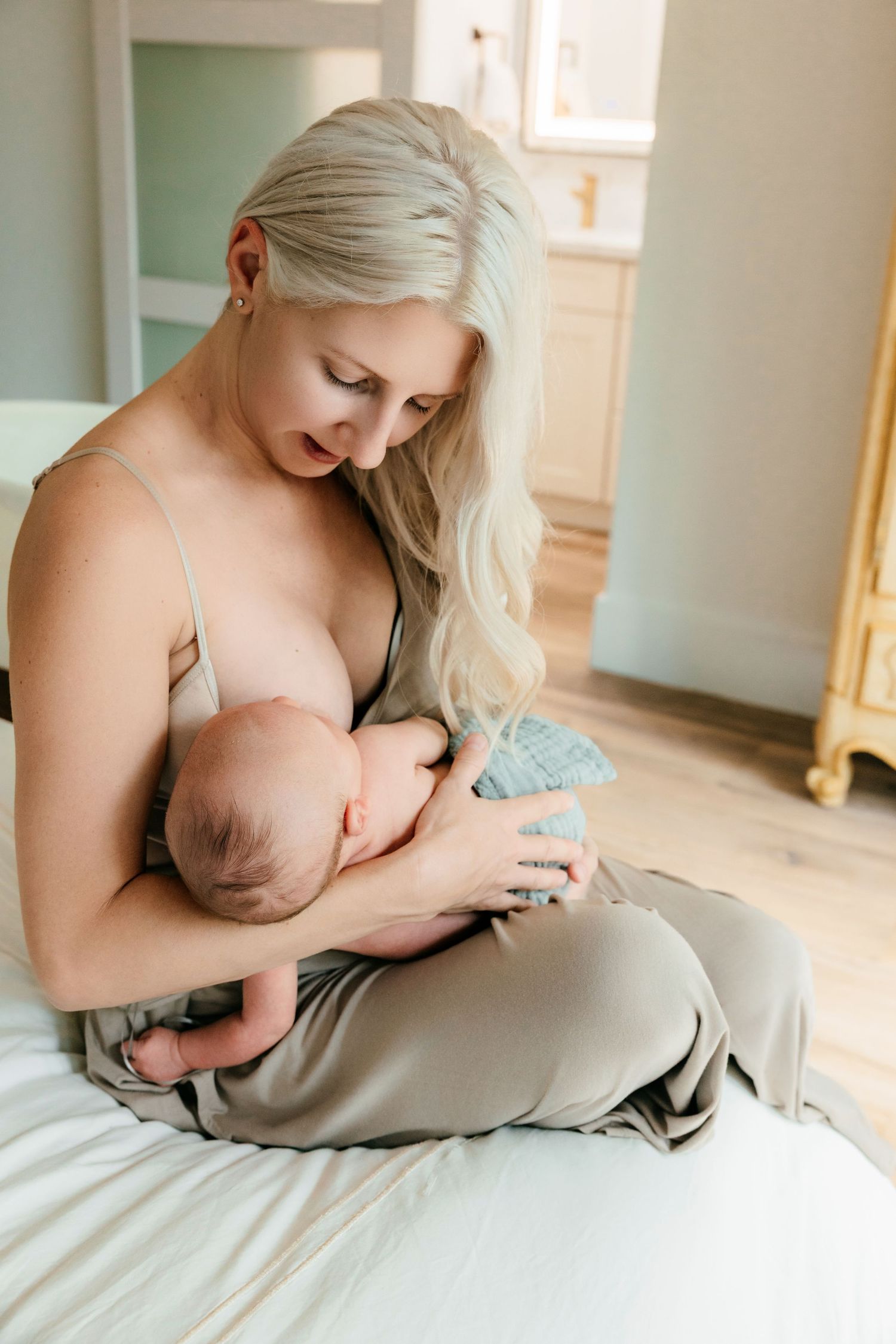 Blonde mother breastfeeding newborn baby while sitting on bed in bright nursery room.