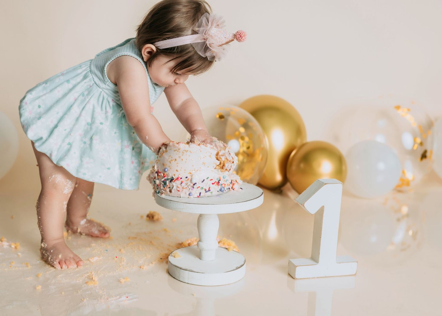Baby girl in blue dress smashing her first birthday cake next to gold balloons and number one decoration.