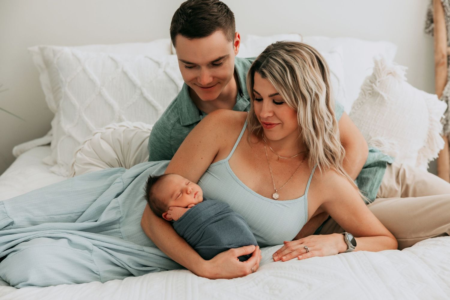 New parents lying on a white bed, lovingly gazing at their swaddled newborn baby wrapped in a teal blanket.