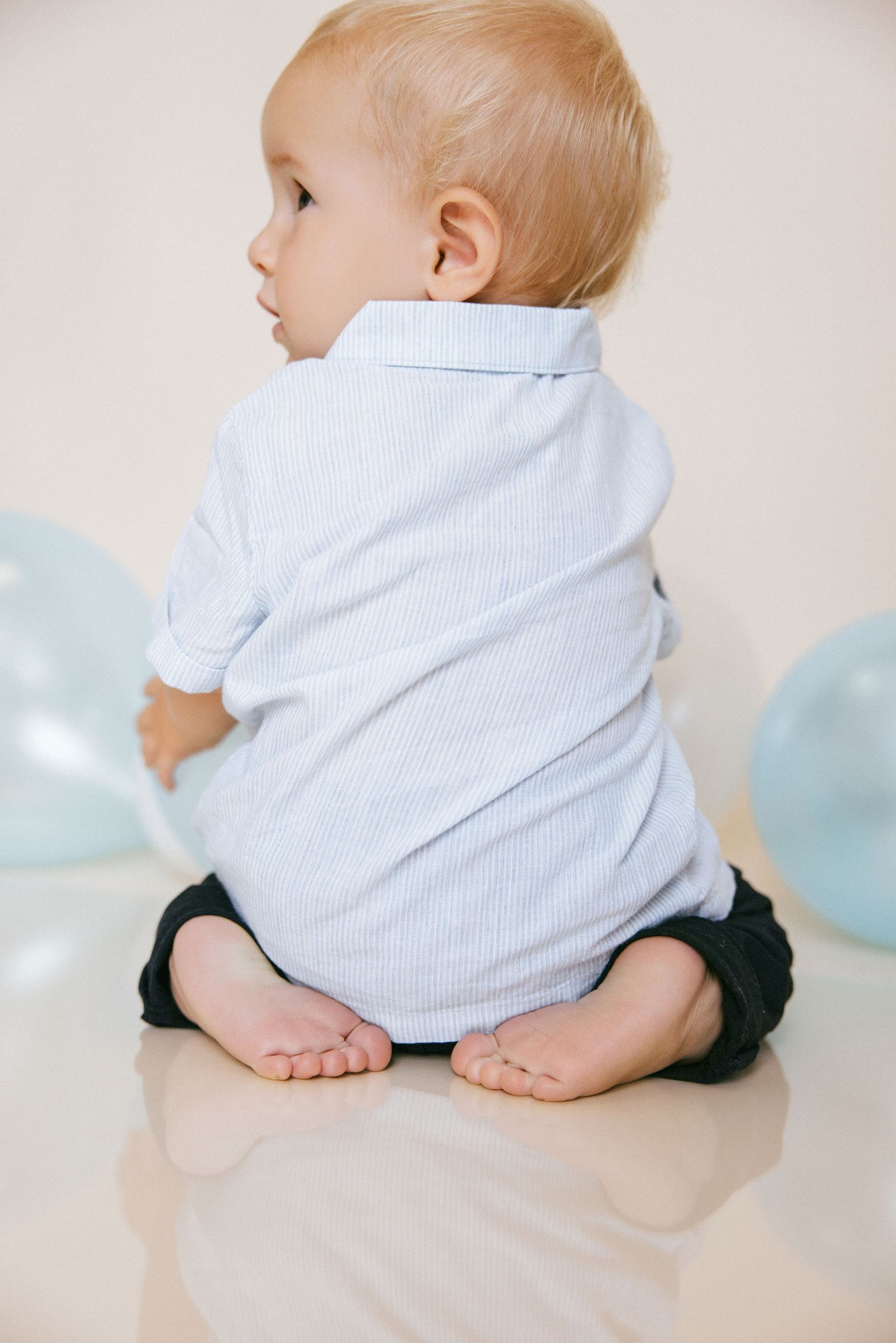 Baby in a striped shirt sitting on floor with blue balloons in background.