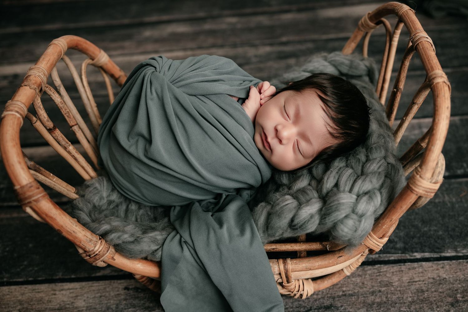Sleeping newborn baby swaddled in green wrap resting in wicker basket with gray knit blanket.