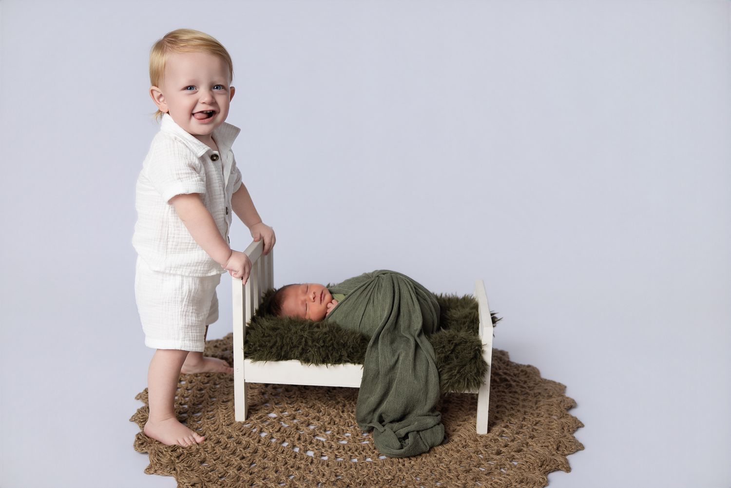 Toddler boy in white outfit stands beside a tiny bed holding a swaddled newborn in green wrap.
