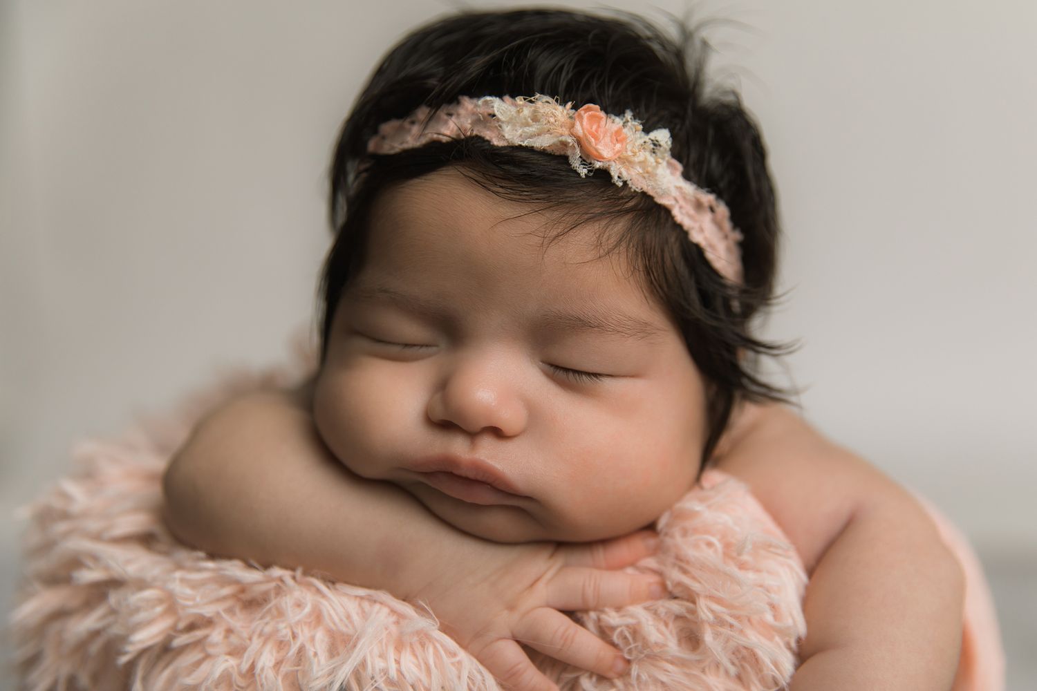 Peaceful newborn baby girl sleeping with dark hair and floral headband, wrapped in soft pink fur blanket.