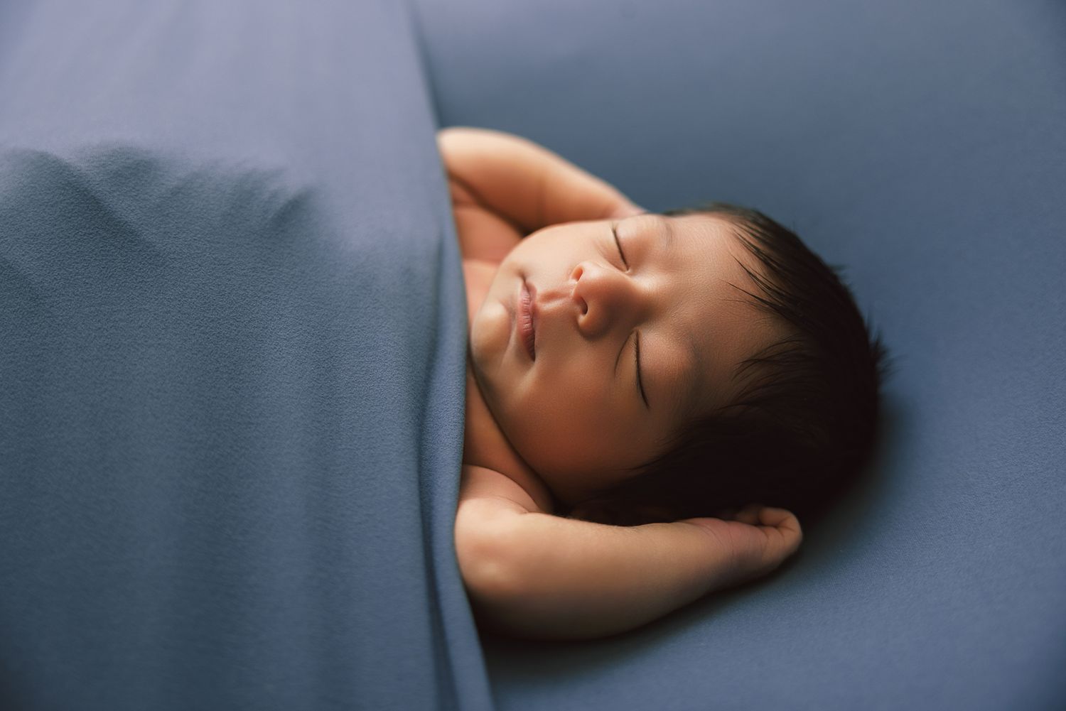 Peaceful newborn baby sleeping on a soft blue blanket, with dark hair and tiny hands visible.