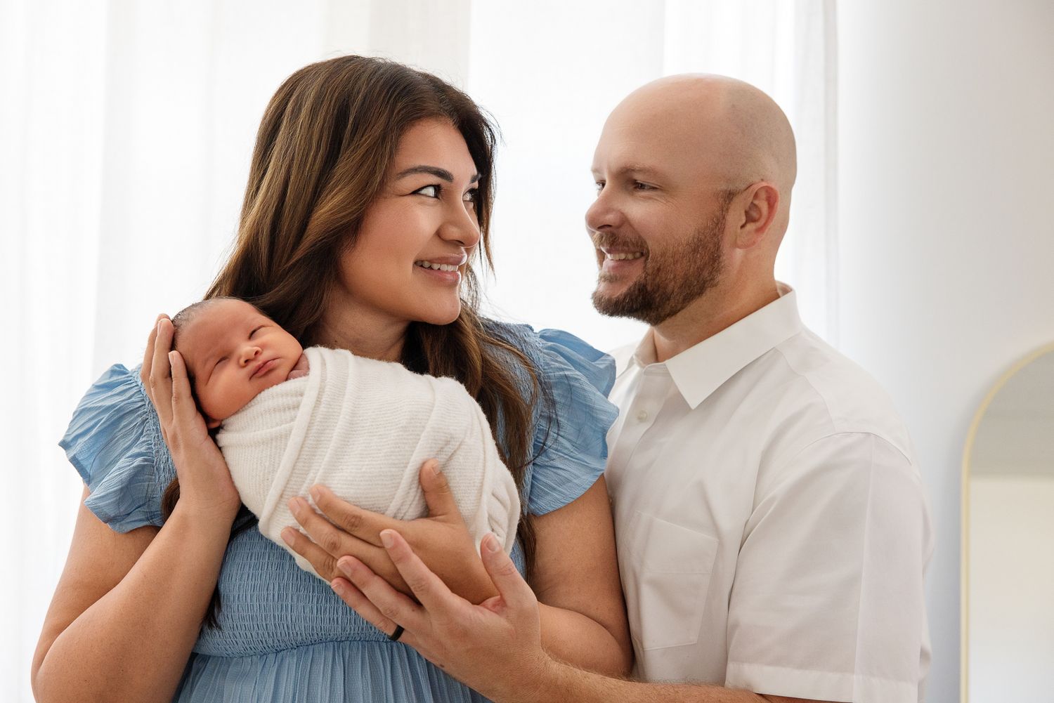 A happy couple smiles at each other while the mother holds their sleeping newborn baby wrapped in white.