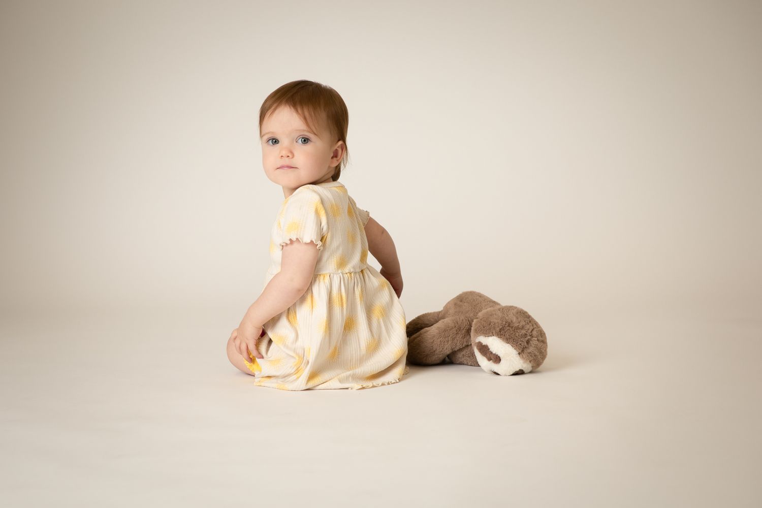 Cute baby girl in yellow striped dress sitting with stuffed sloth toy.