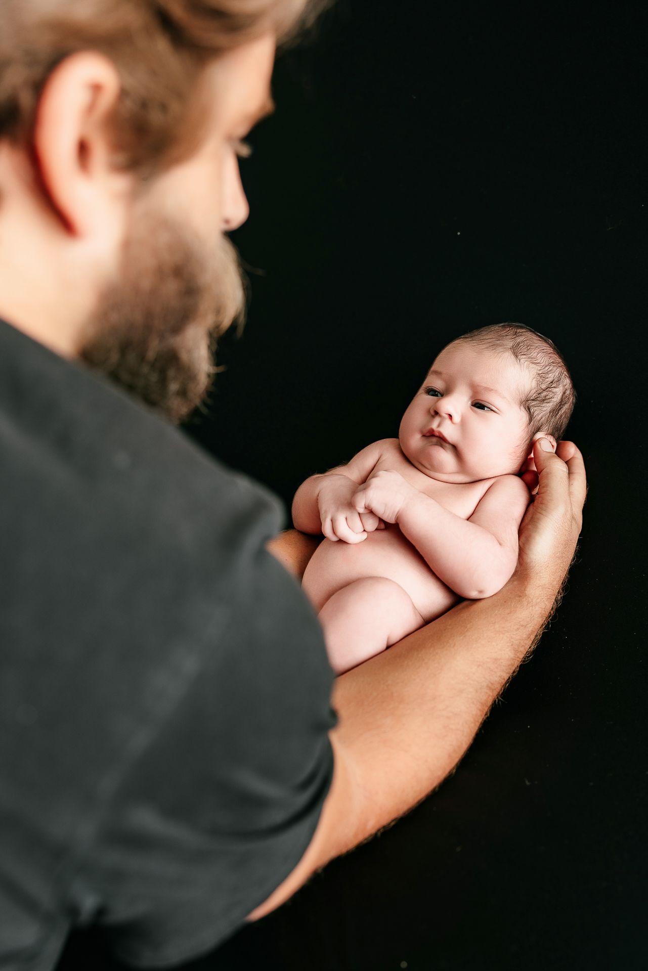 Bearded father tenderly holds naked newborn baby in his arms against a dark background.