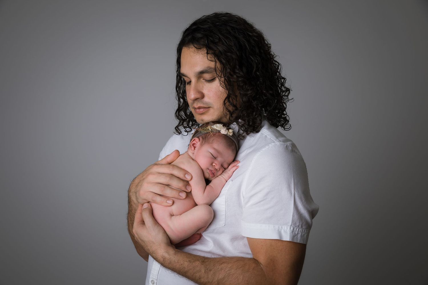 Young father with curly hair tenderly holds sleeping newborn baby against his chest in a studio portrait.