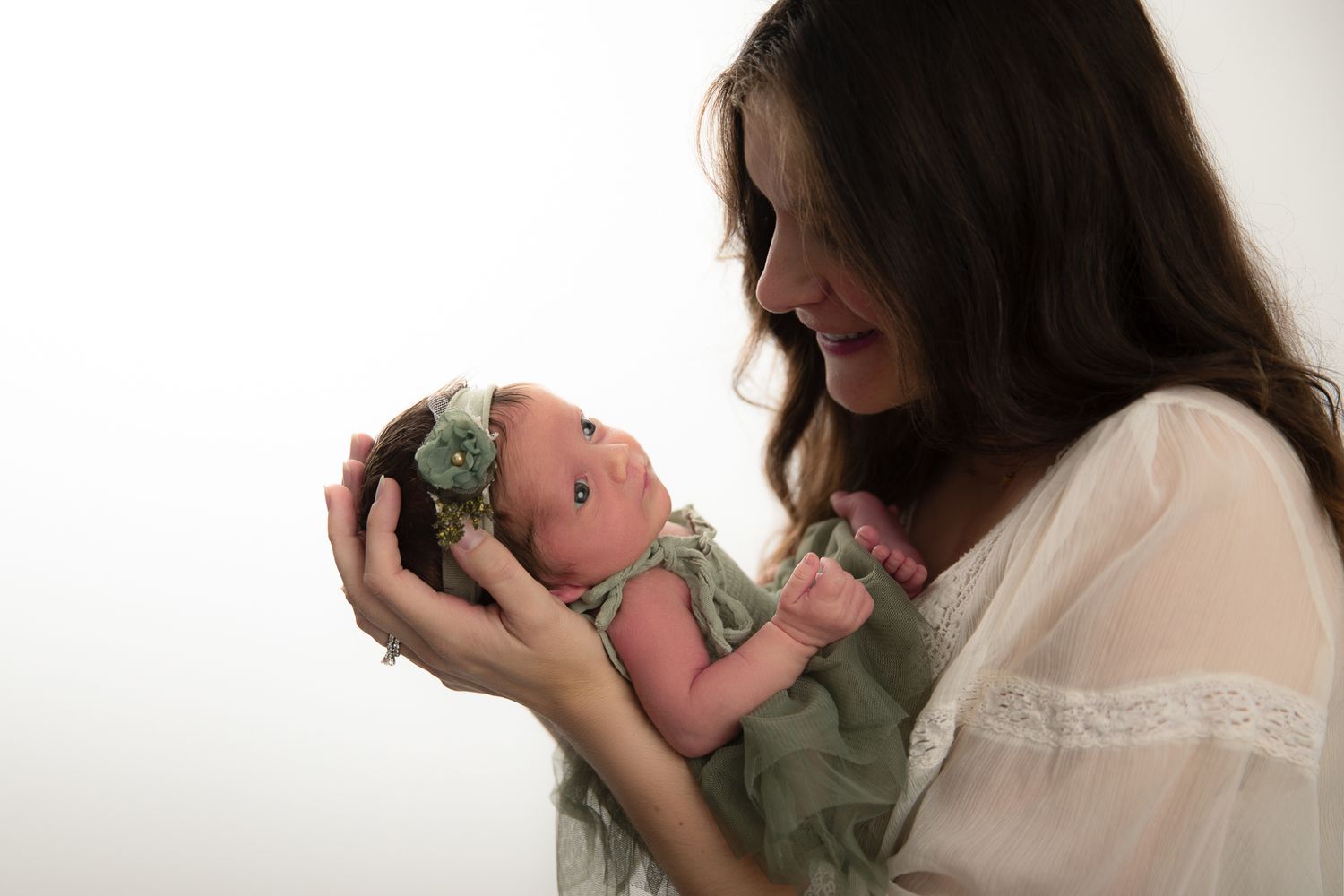 A mother tenderly holds her newborn baby in a green outfit and floral headband during a professional newborn photo session.