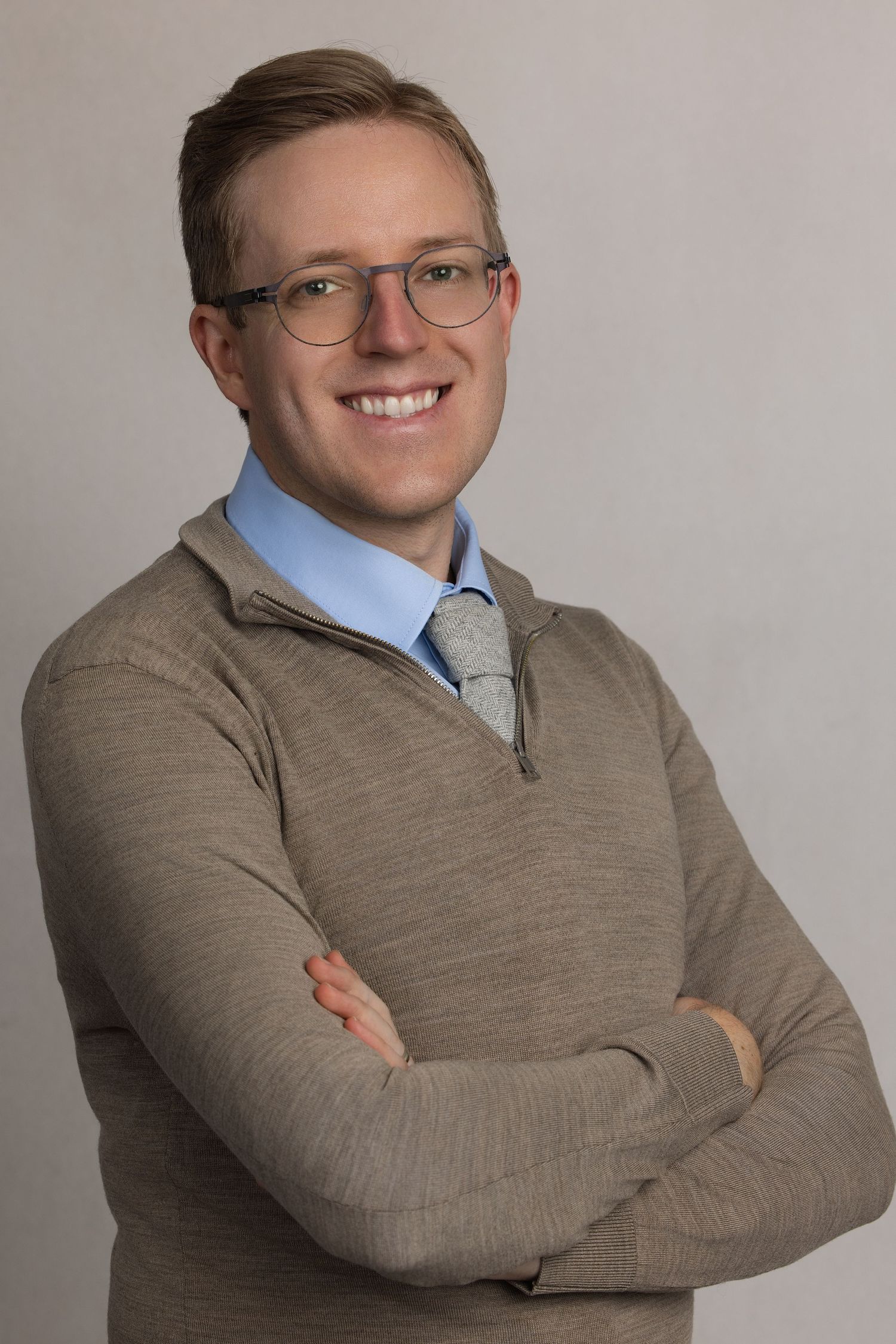 Smiling young professional man with glasses, arms crossed, wearing a gray sweater over blue shirt and tie.
