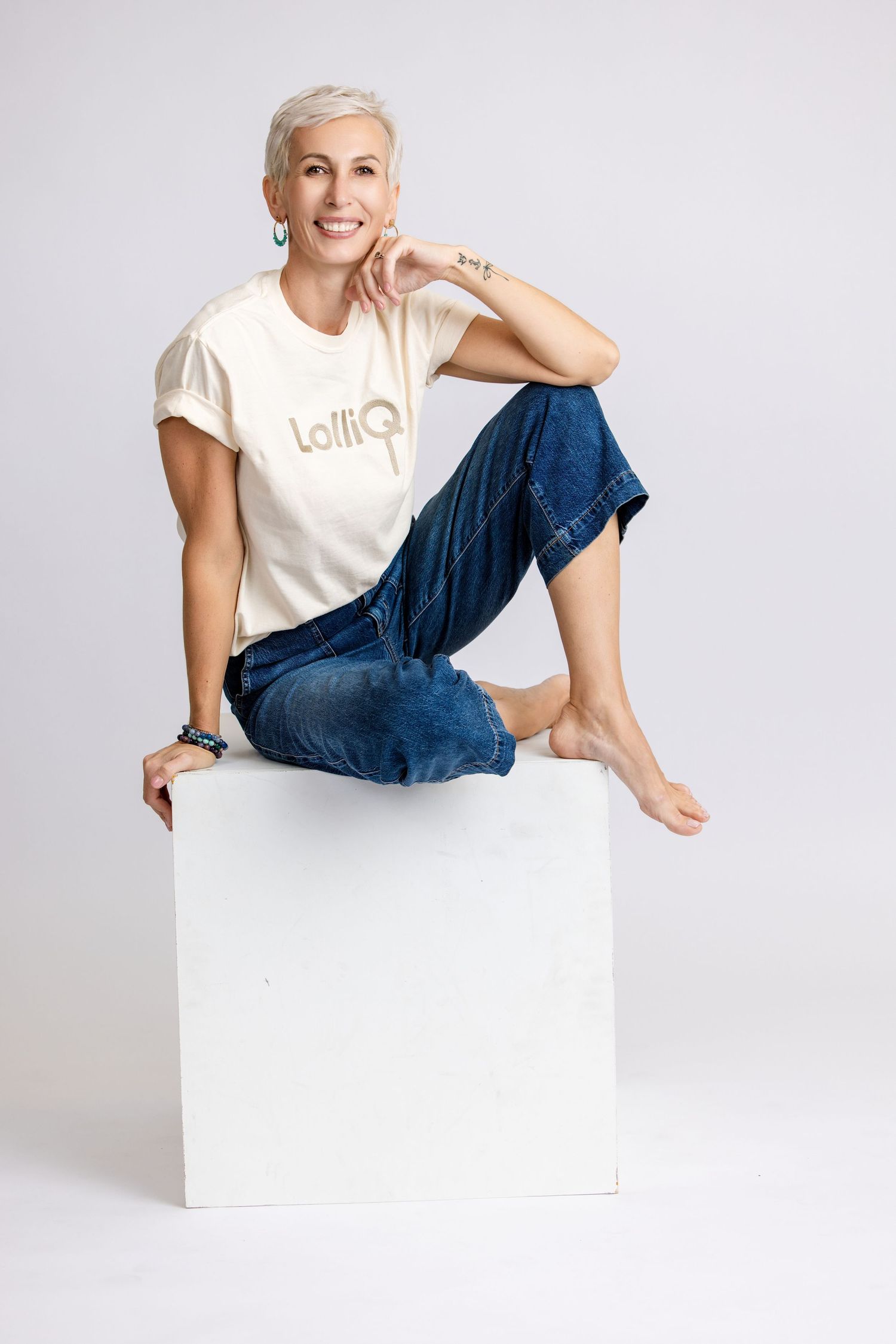 Woman with short gray hair sits barefoot on white cube, wearing cream LolliQ tee and blue jeans, smiling confidently.