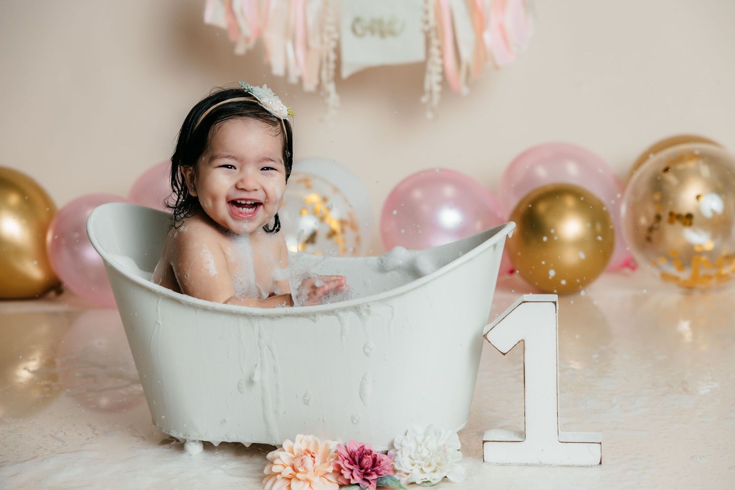 Happy baby girl laughing in white mini bathtub with bubbles, pink and gold balloons, and number one sign.