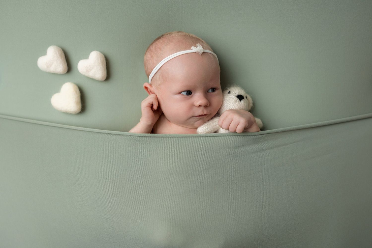 Newborn baby in sage green wrap holding a white teddy bear, surrounded by heart-shaped props.
