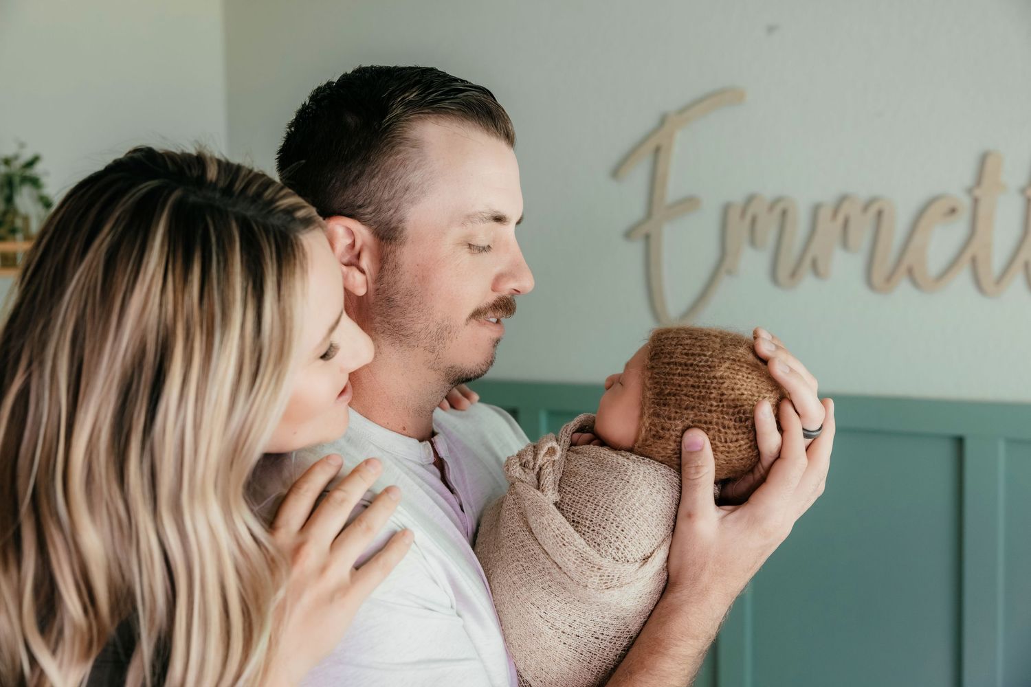 Parents lovingly hold their newborn baby in front of a wooden name sign reading Emmett on a teal wall.