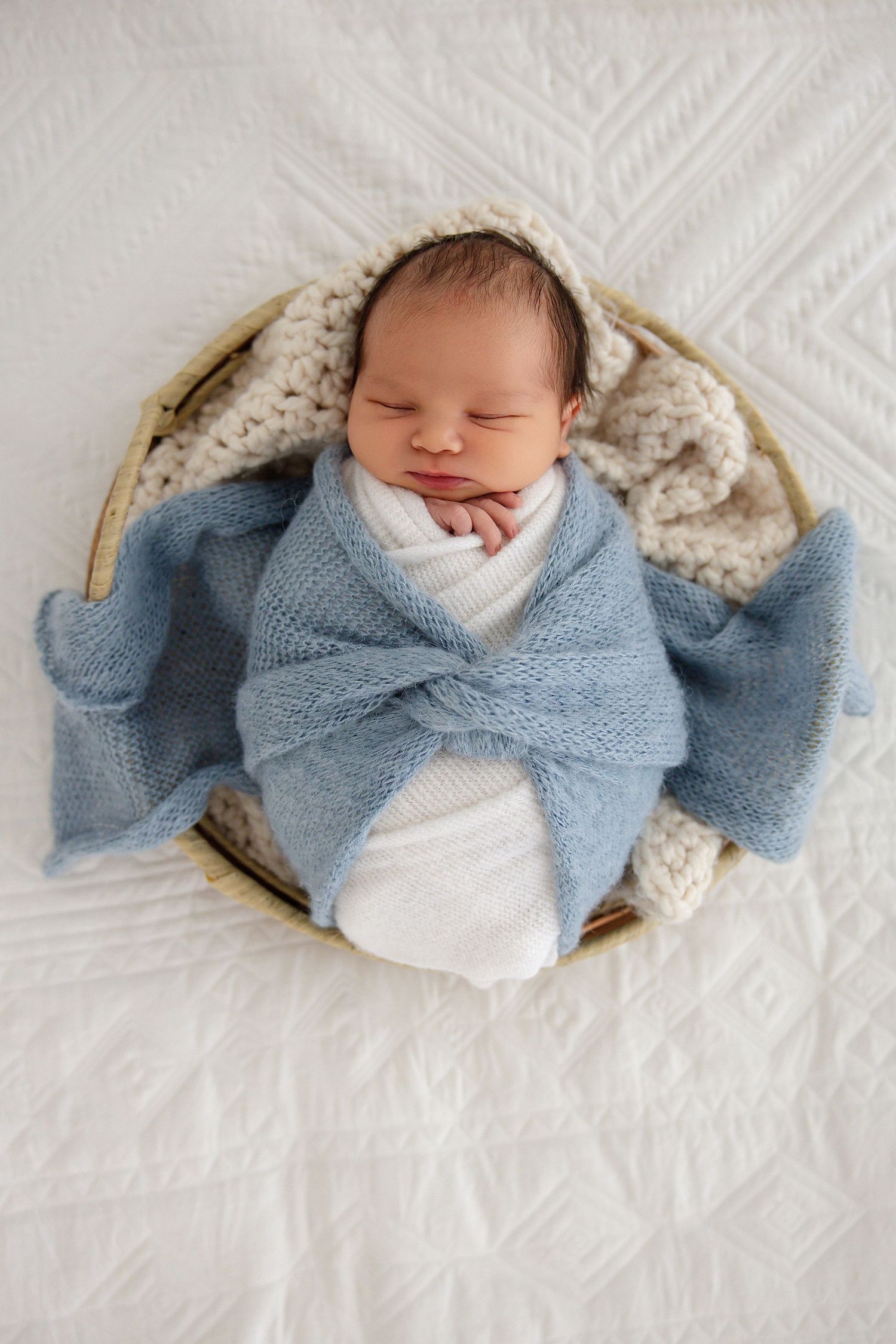 Sleeping newborn baby wrapped in a soft blue knit blanket, nestled in a wicker basket on a white quilted background.