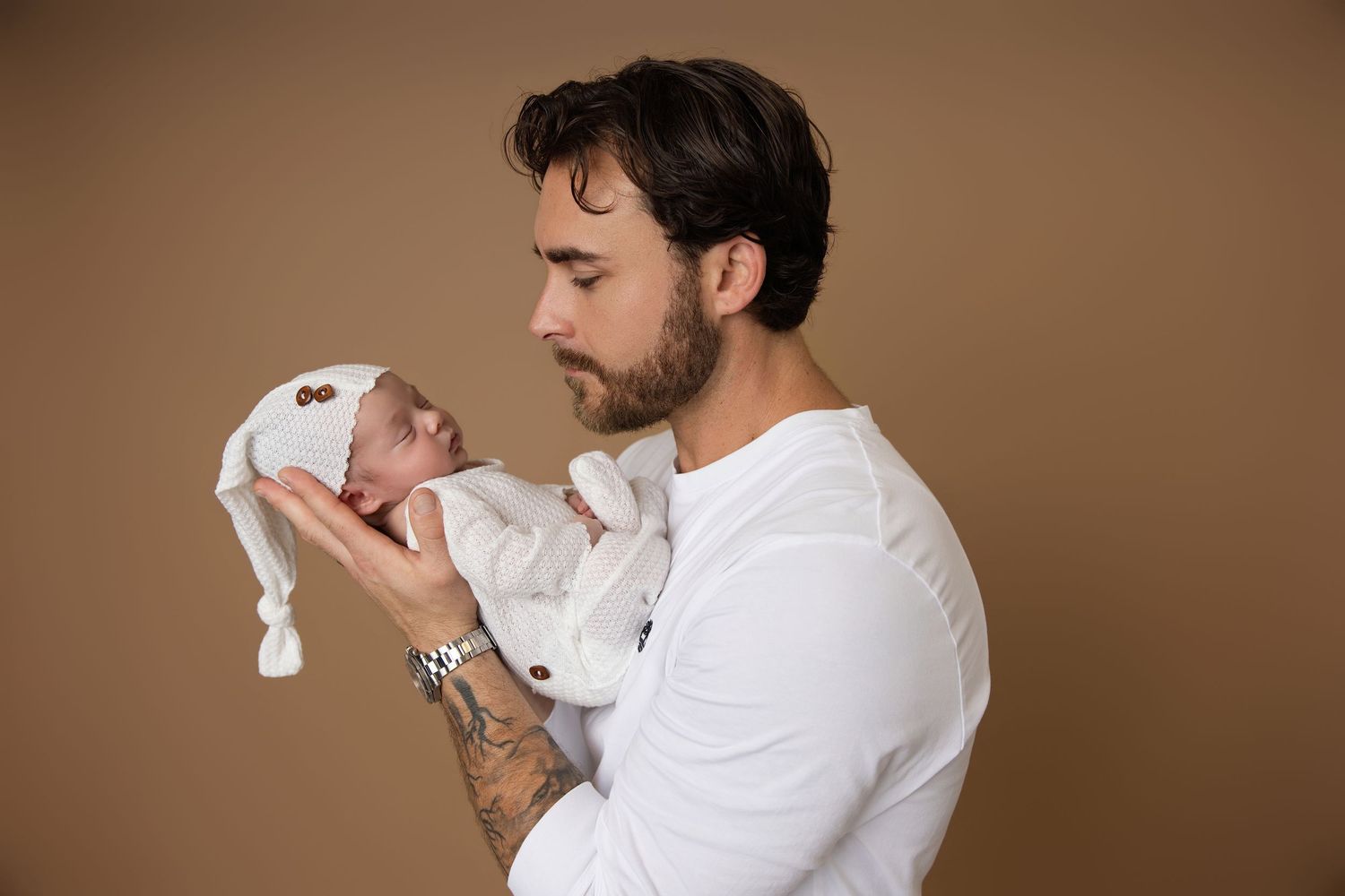 A tattooed father in a white shirt tenderly holds his sleeping newborn baby dressed in a white knit outfit.