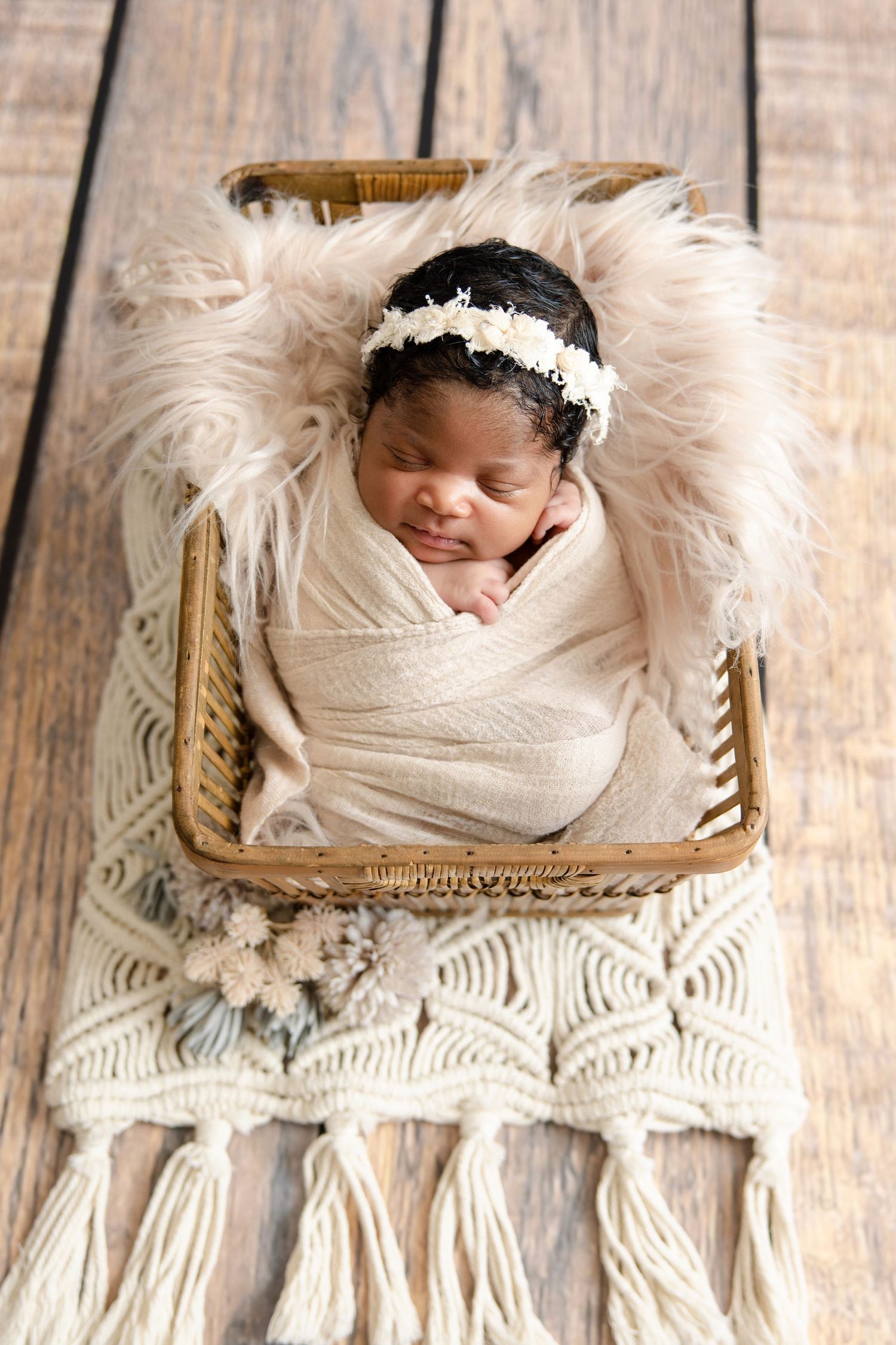 Sleeping newborn baby swaddled in cream wrap with floral headband, resting in wicker basket with fluffy fur lining.