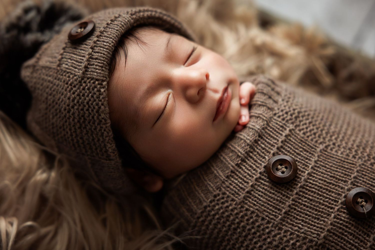 Sleeping newborn baby wrapped in a brown knit swaddle and matching bonnet hat on a soft fur blanket.