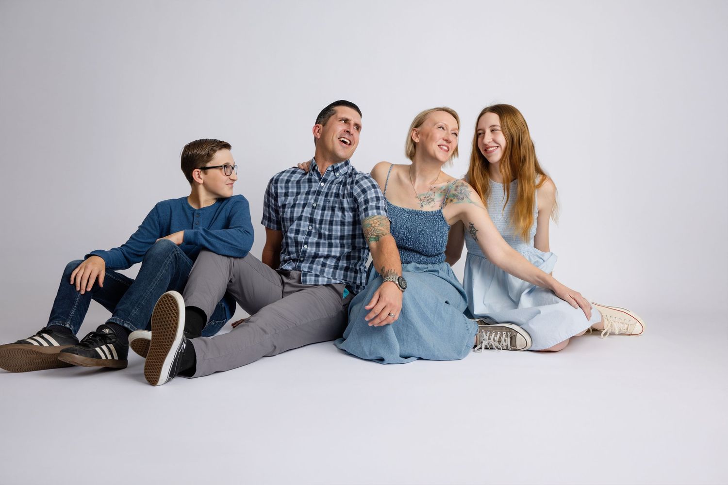 Happy family of four sitting on white floor, laughing and smiling together in casual blue outfits.
