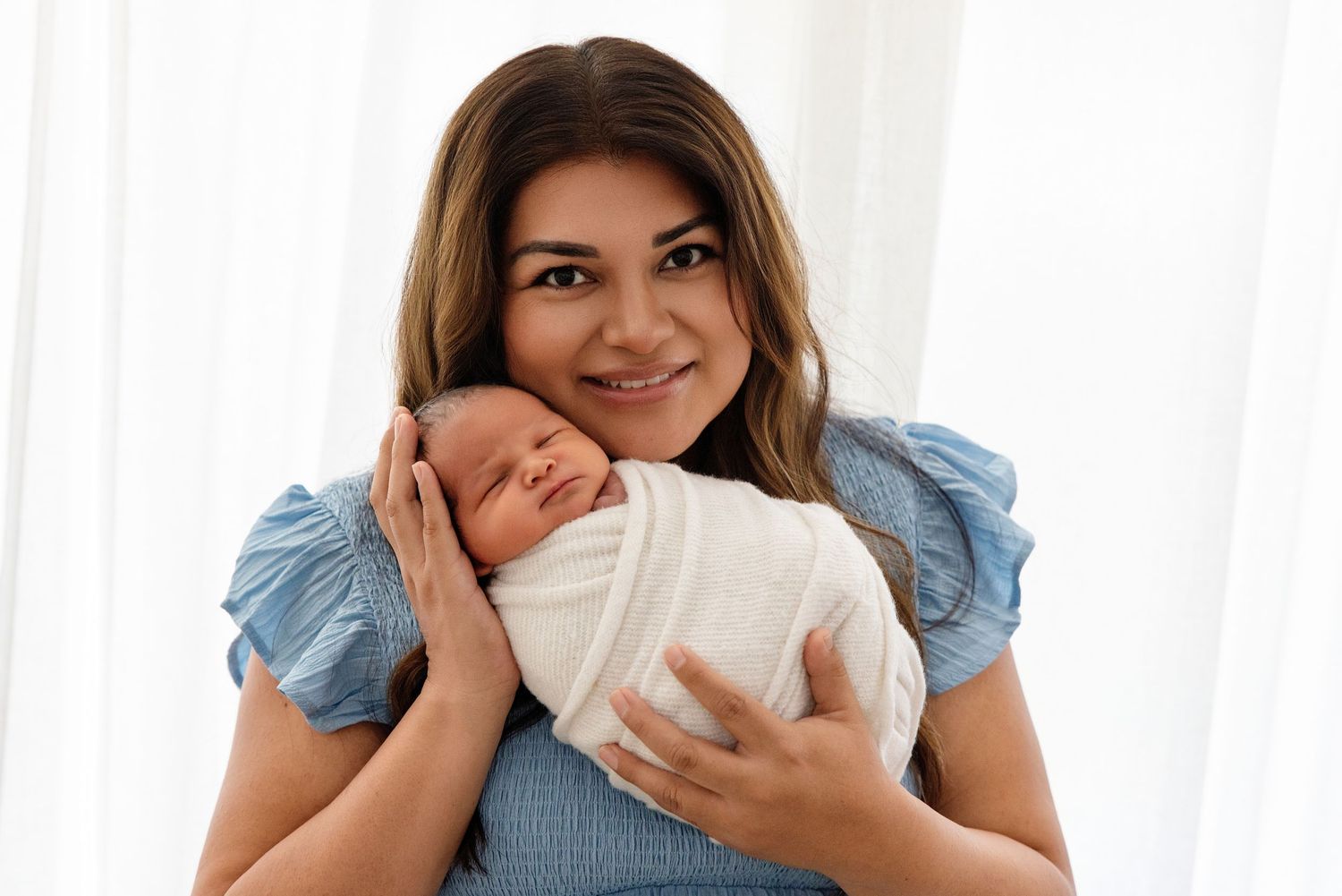 A smiling woman in a blue dress tenderly holds a sleeping newborn baby wrapped in a white swaddle blanket.