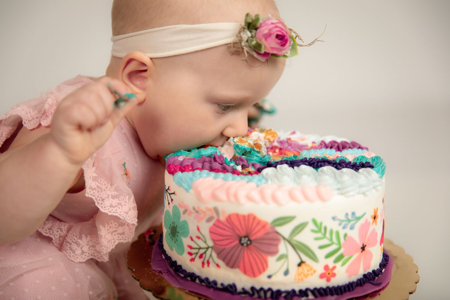 Baby girl in pink dress eating colorful floral birthday cake in studio portrait.