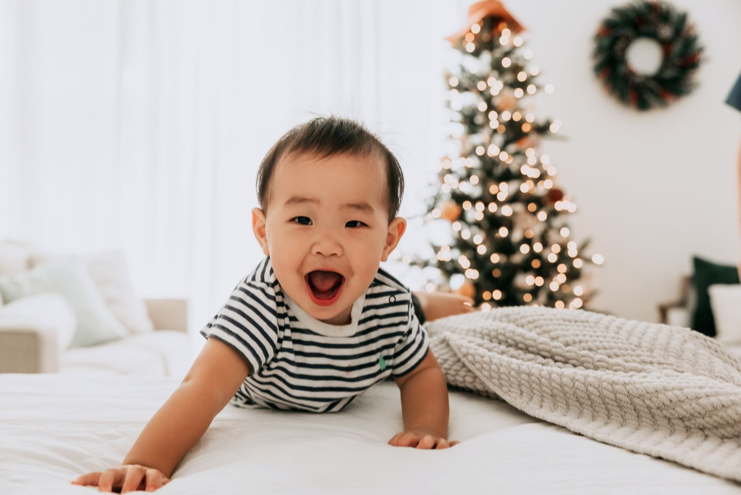 Happy Asian baby in striped onesie crawling on bed near Christmas tree.