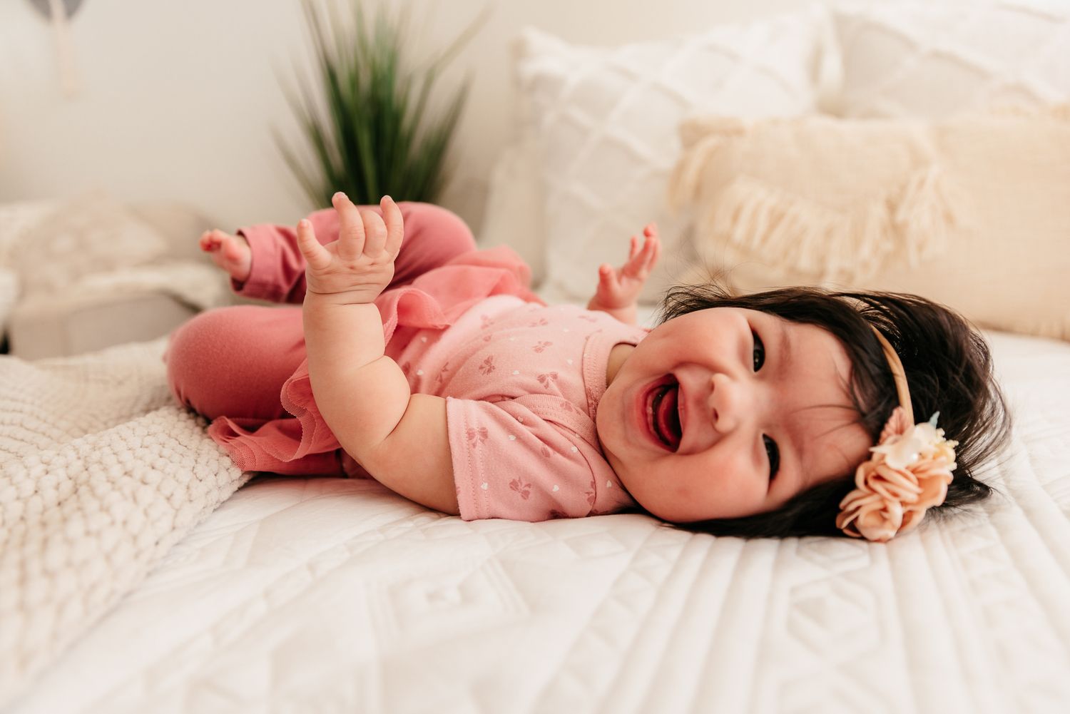 Happy baby girl in pink outfit laughing while lying on a white textured bed with floral headband.