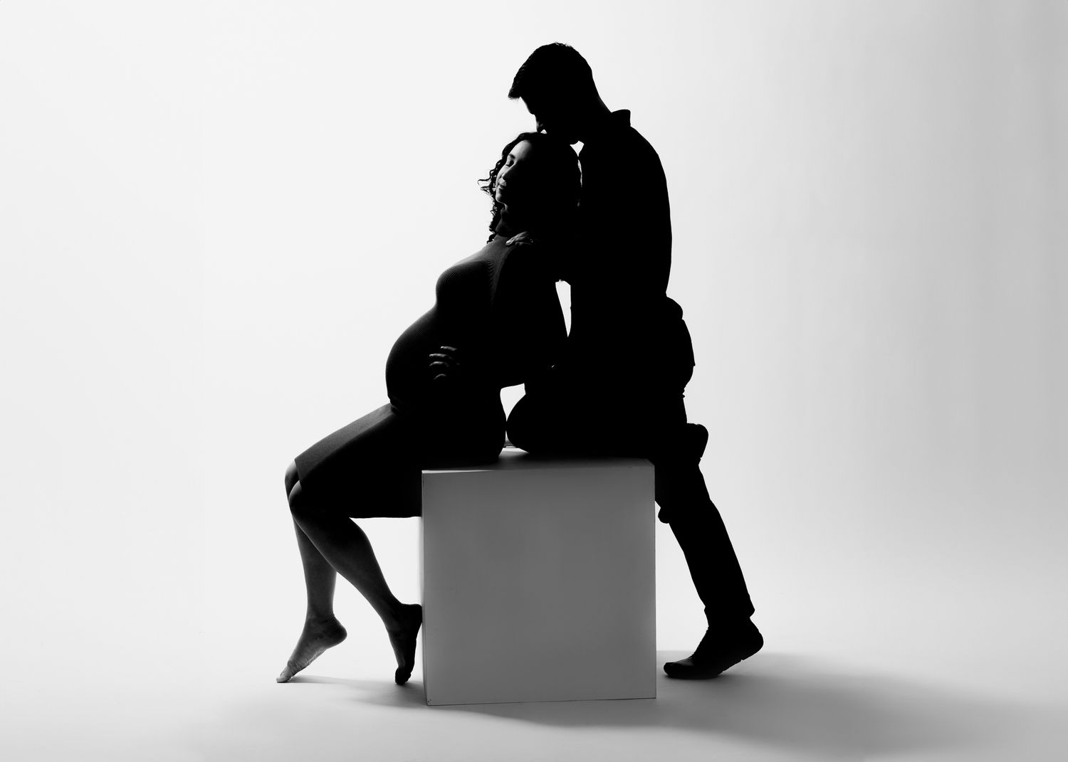 A black and white silhouette showing a couple sitting down on a cube. the man is kissing his pregnant wifes forehead in the Bonsall Studio n