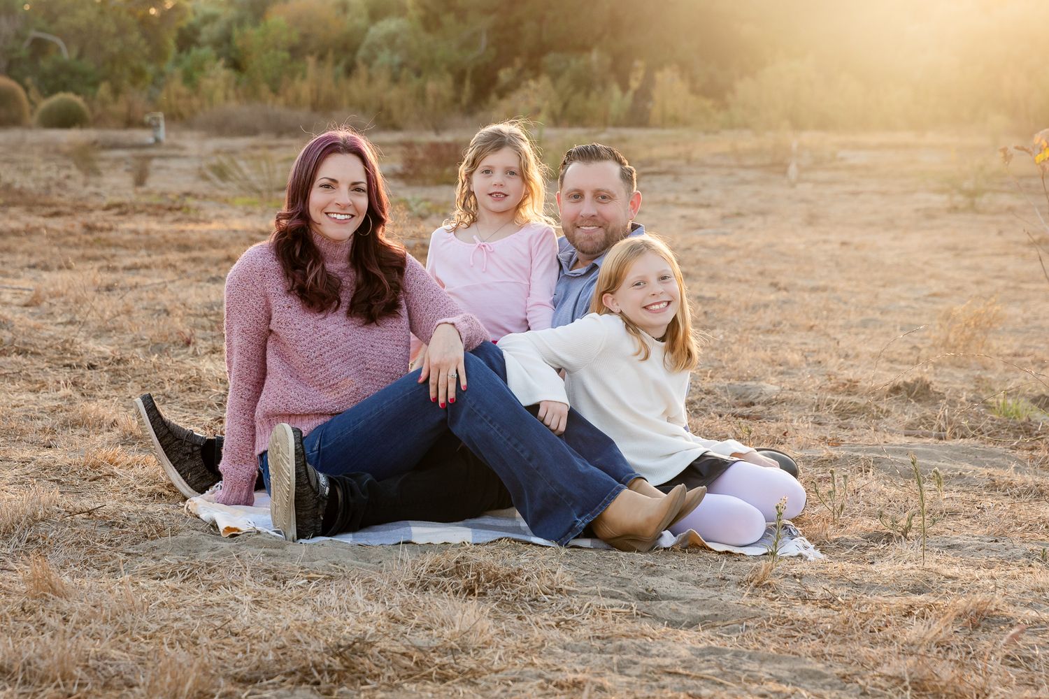 Happy family of four sitting together on blanket in sun-drenched golden field at sunset.