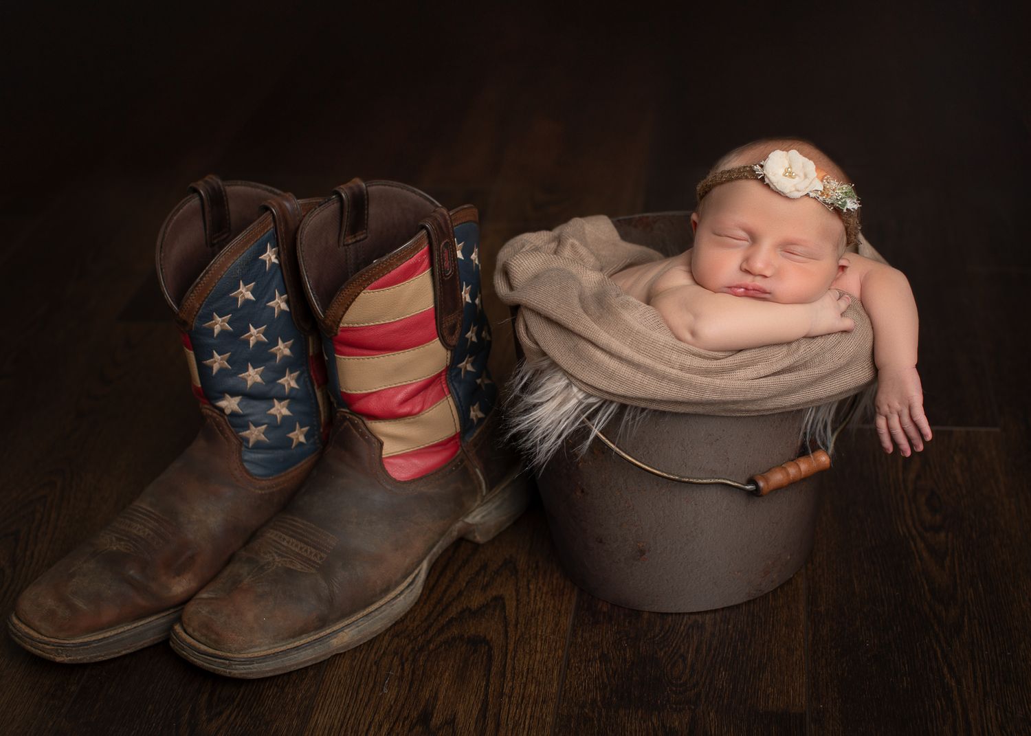 Sleeping newborn baby in a metal bucket beside American flag cowboy boots on dark wood floor.