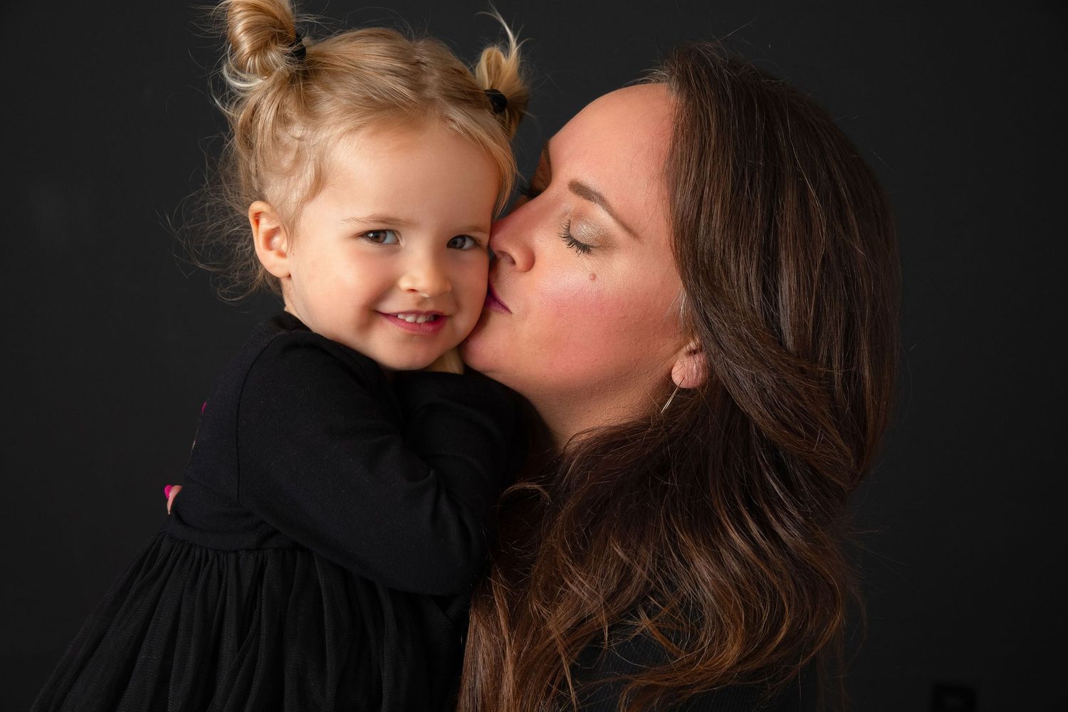 Mother kissing the cheek of her smiling toddler daughter against a dark studio background in the Bonsall Studio