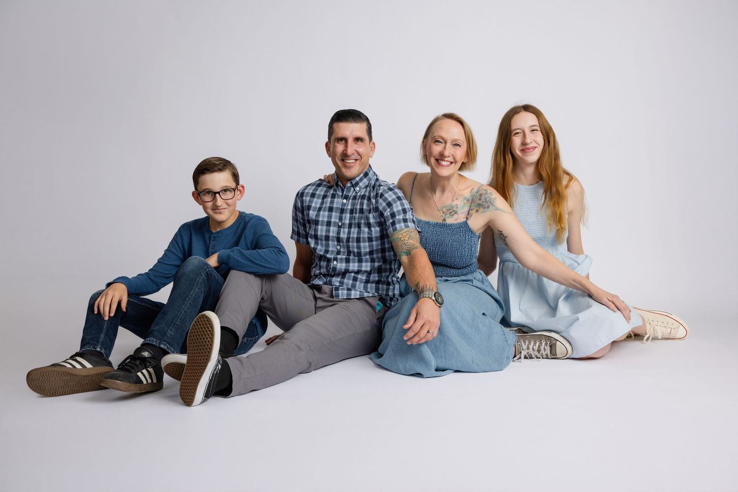 Happy family of four in coordinating blue outfits sitting together on white studio floor smiling.
