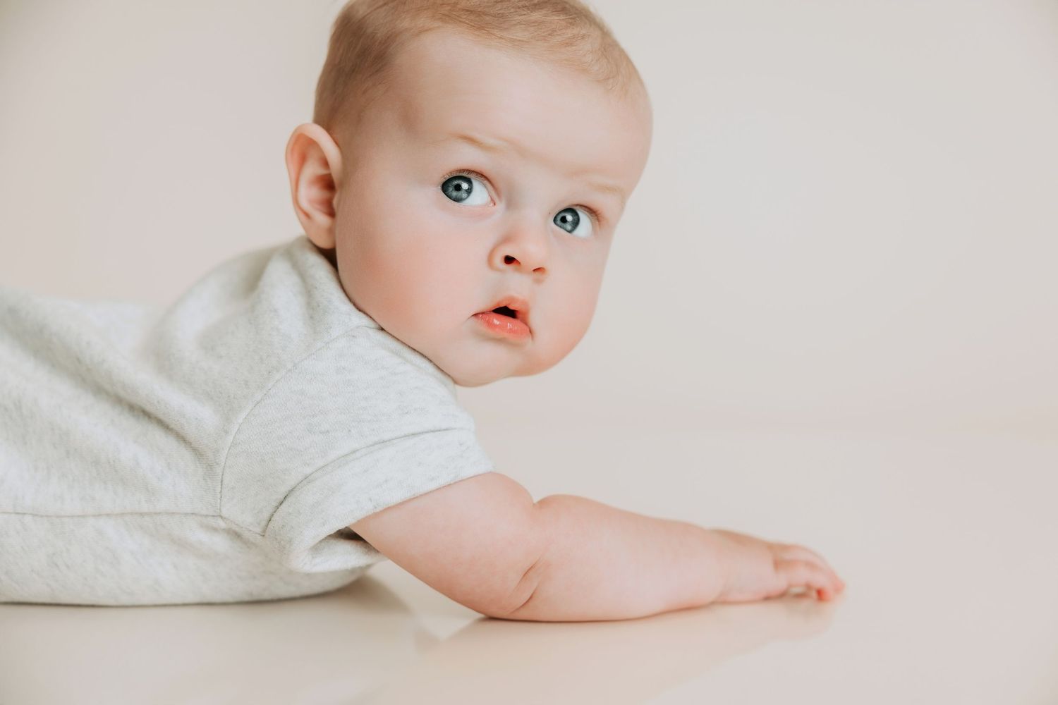 Adorable baby with blue eyes lying on white surface wearing a gray onesie, looking curiously to the side.