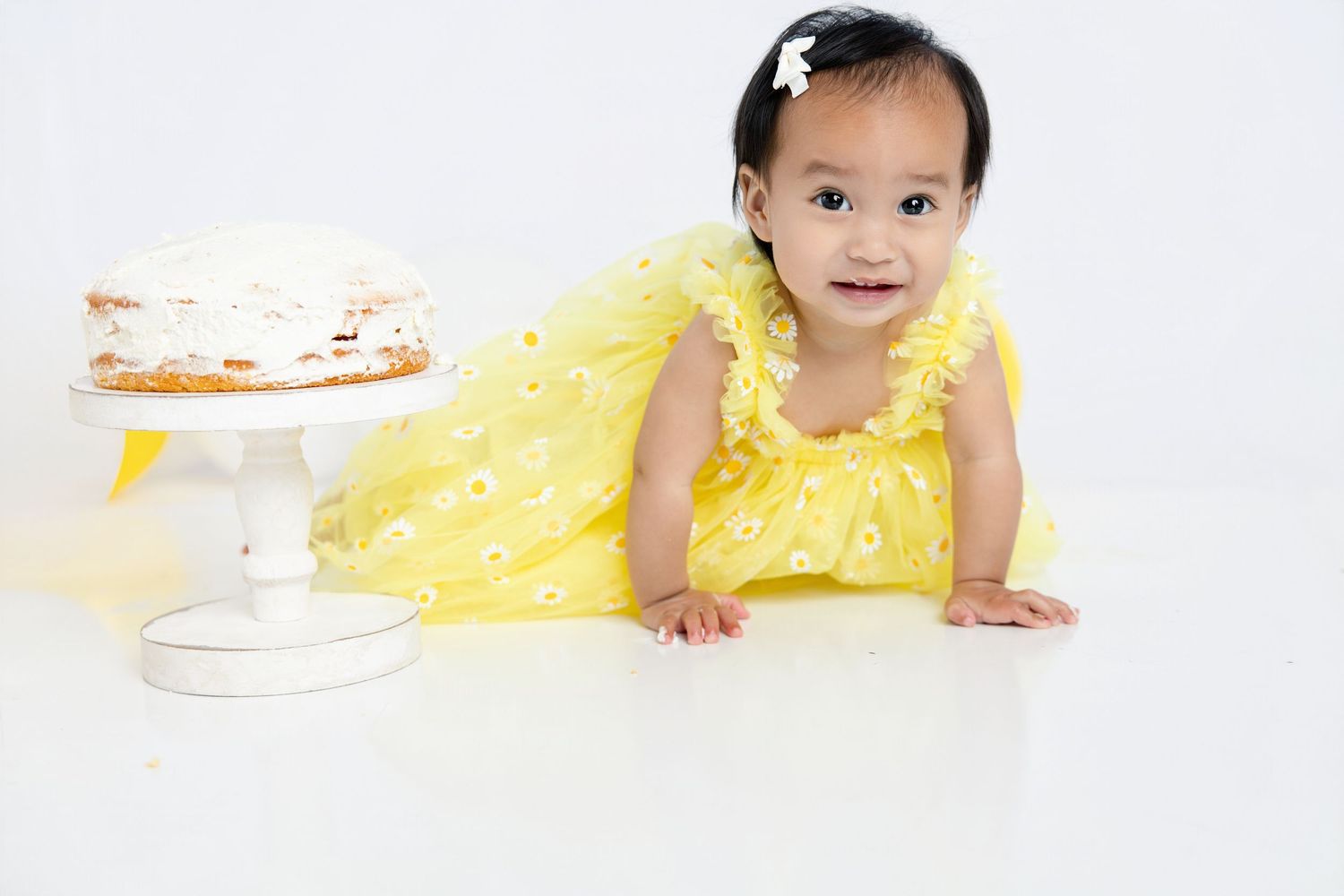 Baby girl in yellow dress smiles near white cake on stand, perfect for first birthday cake smash photography.