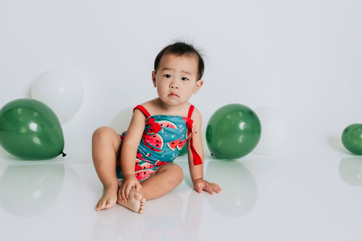 Baby in watermelon romper sits on white floor surrounded by green and white balloons.