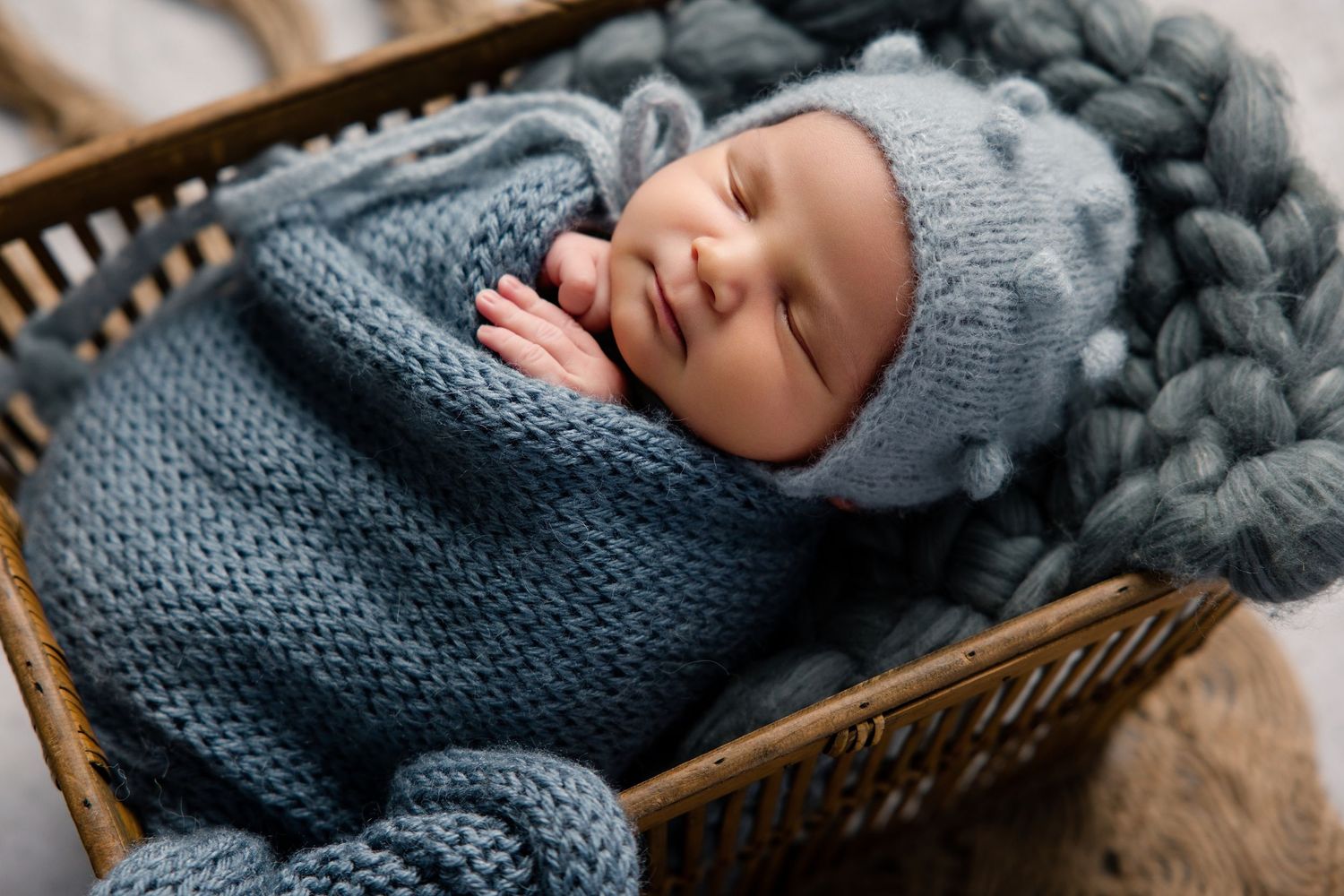 Sleeping newborn baby wrapped in a blue knit swaddle and matching hat, resting in a wicker basket.